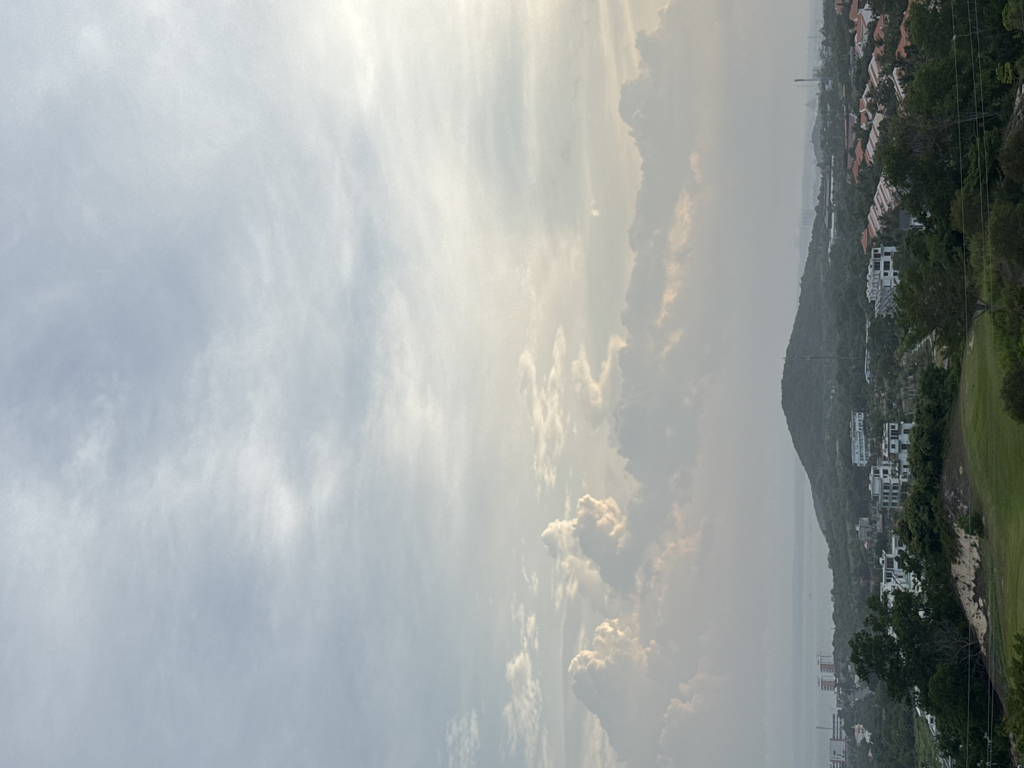The image depicts a scenic view of a coastal town, likely in Phú Thủy, Vietnam, under a cloudy sky with patches of sunlight breaking through. In the foreground, a lush green golf course is visible, with trees and vegetation bordering it. Beyond the golf course, the landscape transitions into a residential area with modern white buildings and houses with orange tile roofs, interspersed with dense greenery and trees. A prominent, rounded hill rises in the middle ground, its slopes covered in trees and vegetation. In the background, the ocean stretches to the horizon, with a few boats visible. The sky is filled with dramatic, textured clouds, some illuminated by sunlight, creating a soft, diffused light over the scene. The overall impression is one of a tranquil, developed coastal environment.