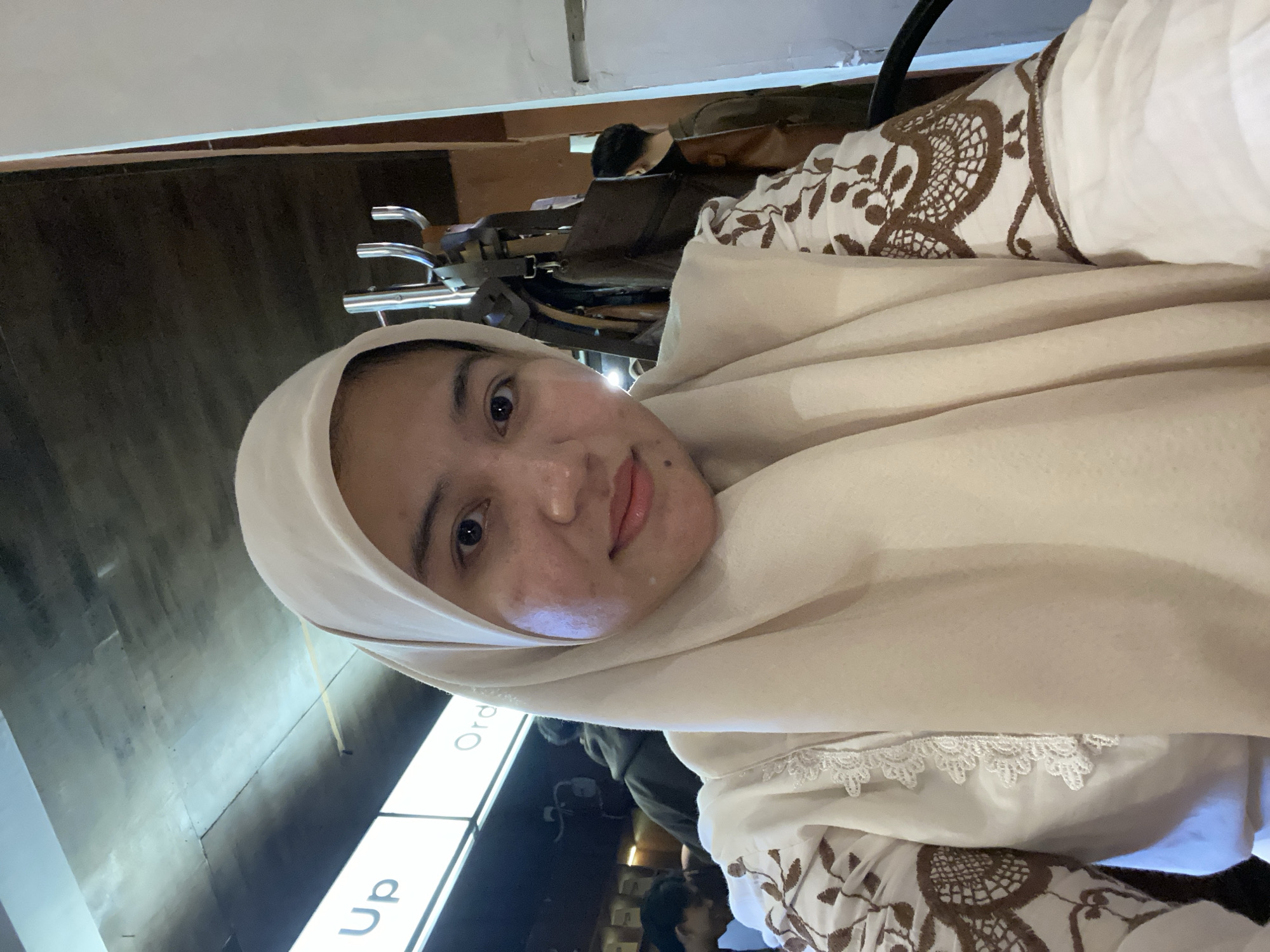 A young woman wearing a light-colored hijab and an embroidered top is taking a selfie indoors. She has brown eyes and a light complexion. She is looking directly at the camera with a slight smile. The background shows a dimly lit interior with a sign that says "Up" and "Ord." Several people are visible in the background, some of whom appear to be waiting in line or ordering food. There are also several bags hanging on a rack. The overall impression is that of a casual dining establishment or a public space where people are ordering services. The lighting is somewhat dim, with a cool-toned light source illuminating the ceiling. The location appears to be a public place in Banjarbaru, Indonesia.