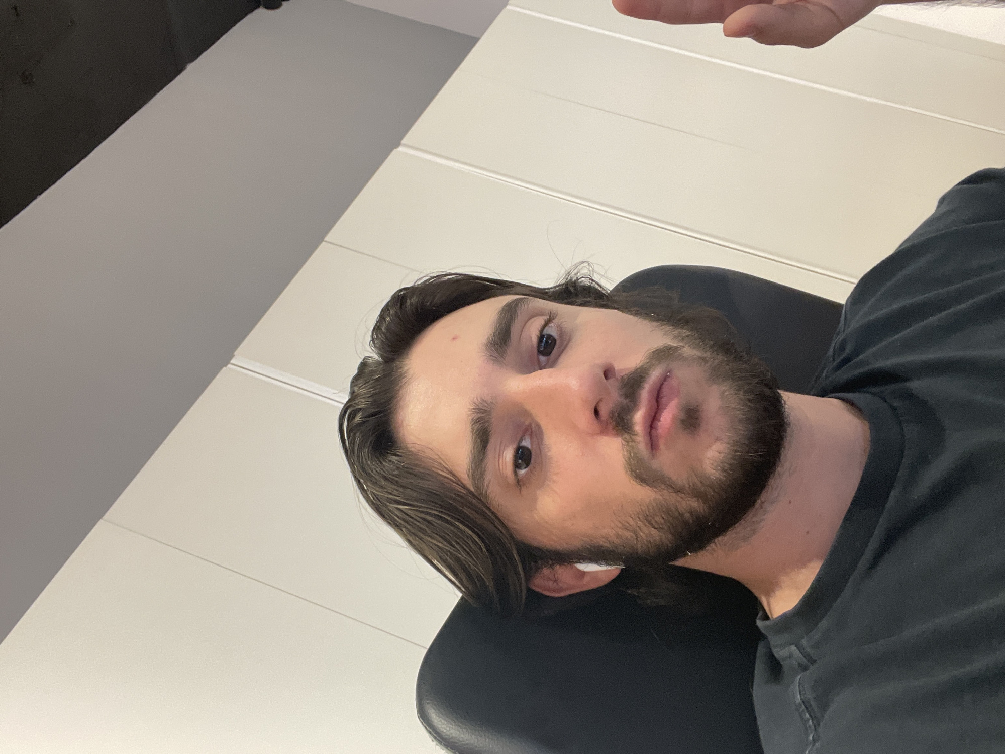 A man with dark hair and a beard is seated in a black chair, wearing a dark gray t-shirt. An earbud is visible in his left ear. The background is composed of a light gray wall. Part of a hand is seen in the upper right corner of the image. The location cannot be determined from the image alone, nor can the specific city in Mexico be identified.