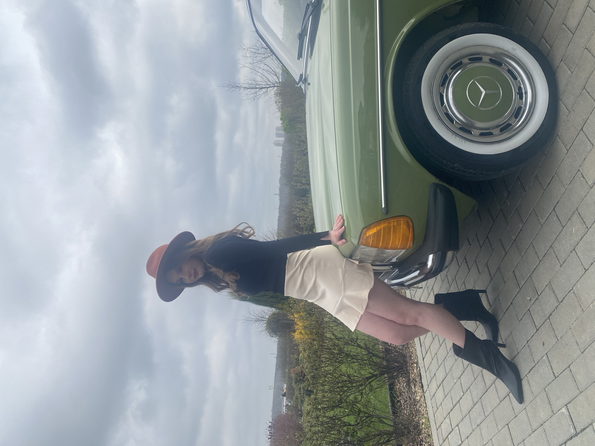 A young woman in a brown hat, black long-sleeve shirt, and cream skirt poses next to a vintage green Mercedes-Benz on a paved surface, likely a driveway or parking area. She leans slightly against the car, resting her hands on its hood. She is wearing black heeled boots. The background includes trees and foliage, with a cityscape faintly visible in the distance. The sky is overcast. The photograph was taken in the city of Yekaterinburg, Russia.