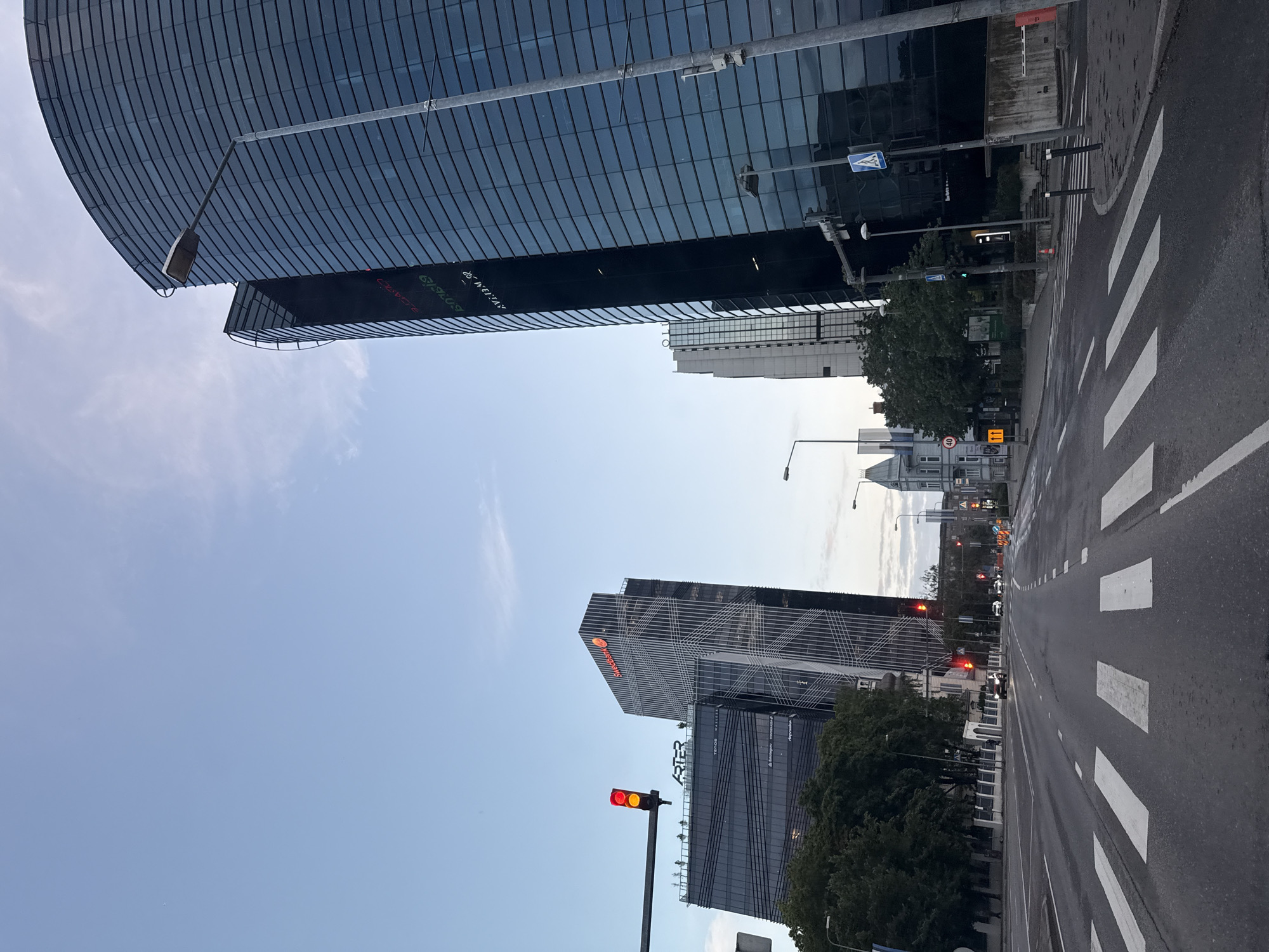 The image shows a view of the city of Tallinn, Estonia, looking down a wide, multi-lane road with pedestrian crossings. On the right side of the road, a tall, modern, curved glass building dominates the foreground. Several other modern high-rise buildings with dark glass facades are visible in the background, along with some older, more traditional buildings. Traffic lights are visible at an intersection, with one showing a red and yellow light. A few cars are on the road, and the sky is a clear blue with some wispy clouds. The overall scene depicts an urban environment with a mix of contemporary and older architecture. The buildings on the right display advertisements or corporate logos, including "SWEDBANK" and what appears to be stock market ticker information. The presence of these financial institutions suggests this is a business or financial district of the city.