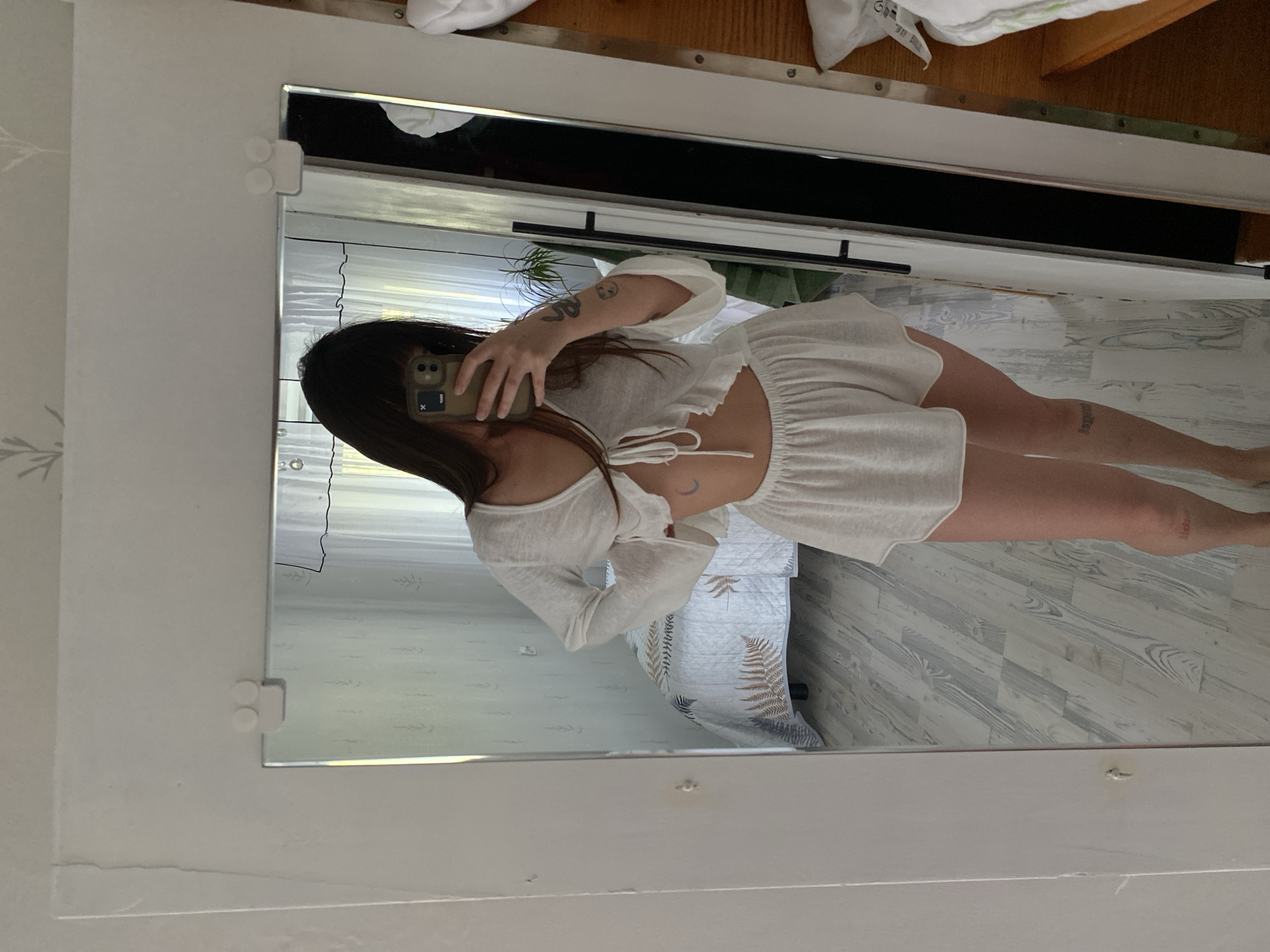 A young woman with long dark hair and bangs is taking a mirror selfie in what appears to be a bedroom in Votkinsk, Russia. She is wearing a white two-piece pajama set, consisting of a tie-front crop top with bell sleeves and high-waisted shorts. A tattoo of a crescent moon is visible on her stomach, and other tattoos are on her arms and legs. The room has light wood-toned flooring and white walls with a subtle pattern. In the reflection, a bed with a patterned duvet is visible, along with a window with white curtains and a plant. The mirror is framed in white and mounted on a white wall, with two white circular mounts visible at the top.