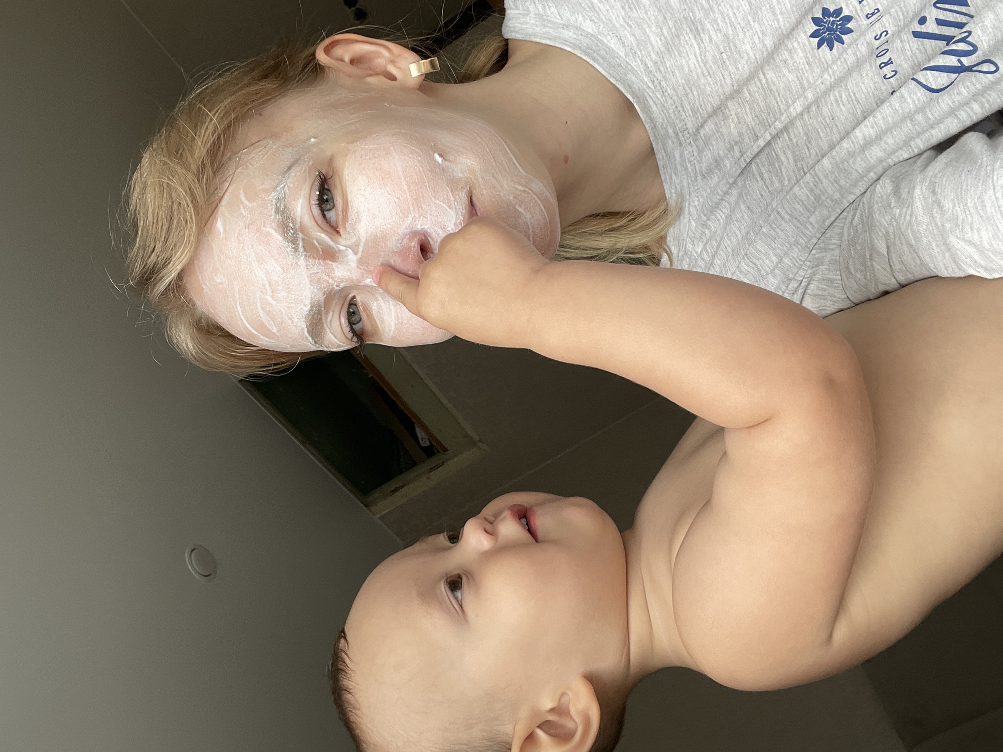A woman with blonde hair and a white face mask is lying on her back. A baby is lying next to her, with their face close to hers. The baby's finger is touching the woman's nose. The woman is wearing a gray shirt with blue writing on it. The ceiling is visible in the background. The city of Perm, Russia, is not identifiable in the image.