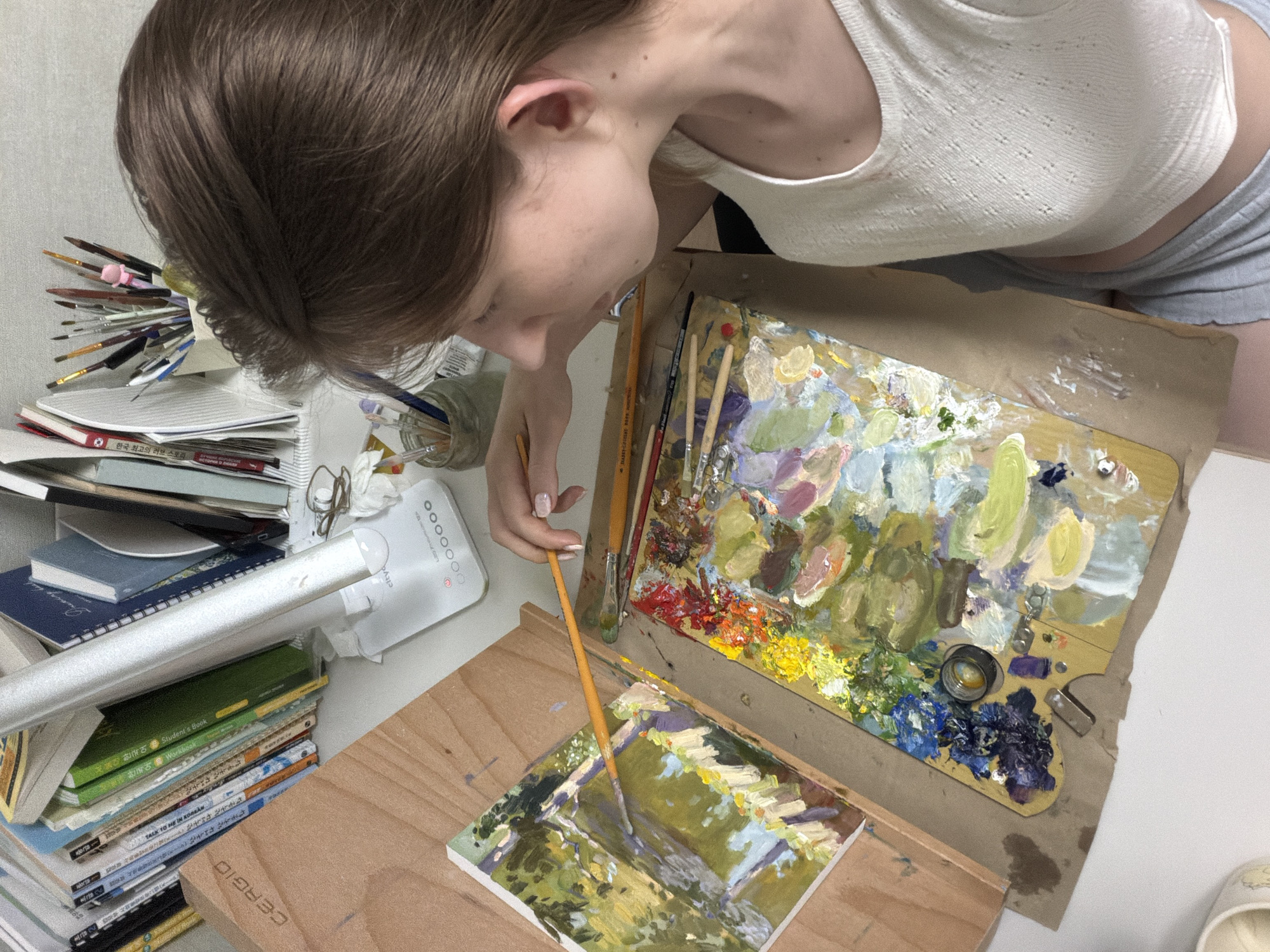 A young woman is painting a landscape outdoors, possibly in a park or garden in Dongducheon-si, South Korea. She is focused on her work, with a palette of colorful paints and brushes laid out before her. The painting, rendered in a loose, impressionistic style, depicts trees and sunlight filtering through the leaves. The woman is wearing a white top and grey shorts. The background shows stacks of books and art supplies, suggesting an indoor setting before moving outdoors to paint.