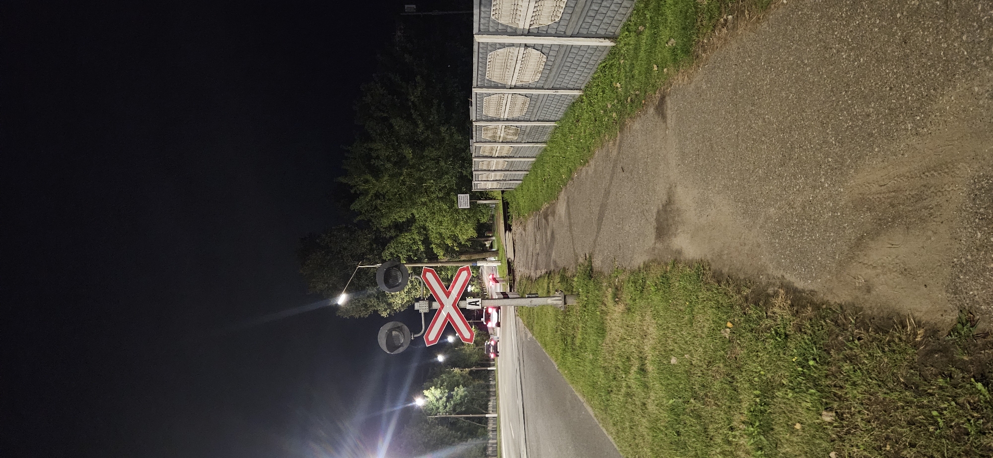 The image captures a nighttime scene at a railroad crossing on an asphalt road in Vitsebsk, Belarus. The foreground features a paved pedestrian walkway and a grassy verge. To the left, a two-lane road carries two vehicles, their headlights illuminating the scene. The center of the image is dominated by a railroad crossing signal, a red and white X-shaped sign beneath two round signal lights. The sign has the letter "A" printed on it. A tall pole supports the signal lights and sign. Beyond the road, trees line the landscape, their dark silhouettes contrasting with the night sky. On the right side of the walkway, a grey concrete fence with patterned inserts runs parallel to the path. The overall impression is one of a quiet, functional area at night, likely a residential or suburban setting given the presence of the fence and sidewalk.