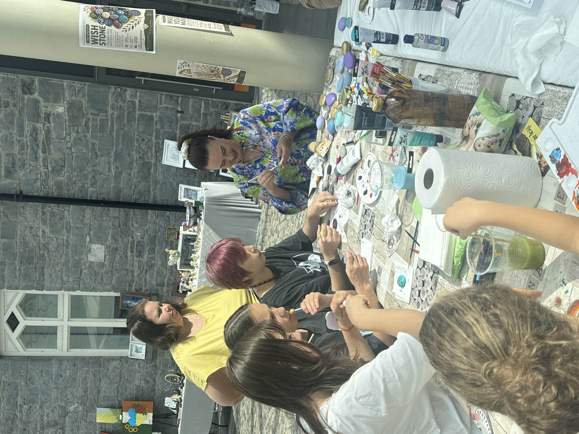 The image shows a group of people at a table, painting rocks outdoors. The table is covered with a patterned cloth and is laden with art supplies including paints, brushes, water cups, and a roll of paper towels. Several rocks, some already painted and others blank, are laid out on the table. There are four individuals clearly visible: a woman in a yellow top, a woman in a floral top, and two younger individuals with dark hair. The woman in the floral top is actively painting a rock. The background reveals a stone building with a large, gothic-style window, suggesting the location might be a historical site. Signs are visible on a pole in the background, one reading "WISH STONE" and another partially visible that says "MAKE YOUR MAR...". Given the context of the "Wish Stone" sign and the stone building, the scene is likely taking place at the Kilkenny Design Centre or a nearby artisan area in Kilkenny city, Ireland, known for its crafts and heritage buildings. The atmosphere appears to be relaxed and creative, possibly an art workshop or a community event.
