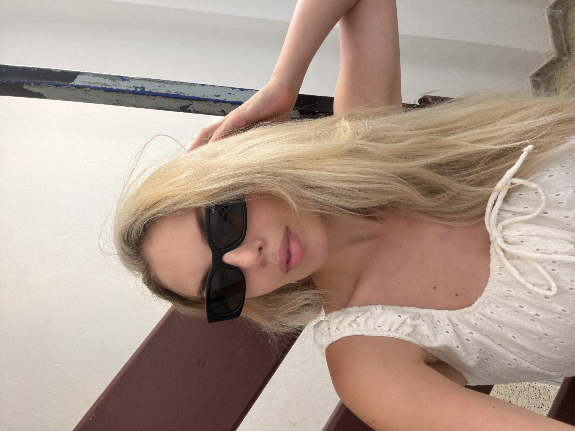 A young blonde woman wearing black sunglasses and a white eyelet top is posing for a selfie. She is leaning against a reddish-brown railing, with a white wall and a blue metal pole in the background. The image appears to be taken on a staircase. The woman has her right hand on the back of her head, and her hair is long and blonde. The overall setting is an urban environment, likely a public space within Benimàmet-beniferri, Spain, suggested by the visible architectural elements typical of the region.