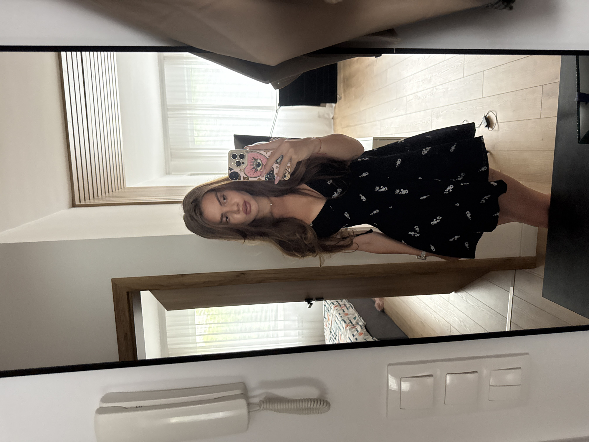 A young woman with long, wavy brown hair is taking a mirror selfie indoors. She is wearing a black, short-sleeved dress with a floral pattern. The dress has a V-neck and a flared skirt. She is holding a pink phone with a heart-shaped case. In the reflection of the mirror, a bedroom is visible with a bed and a window with white curtains. To the left of the woman, there is a white intercom system mounted on the wall, and a light switch panel below it. The floor is made of light-colored wood. The overall scene suggests a domestic setting.