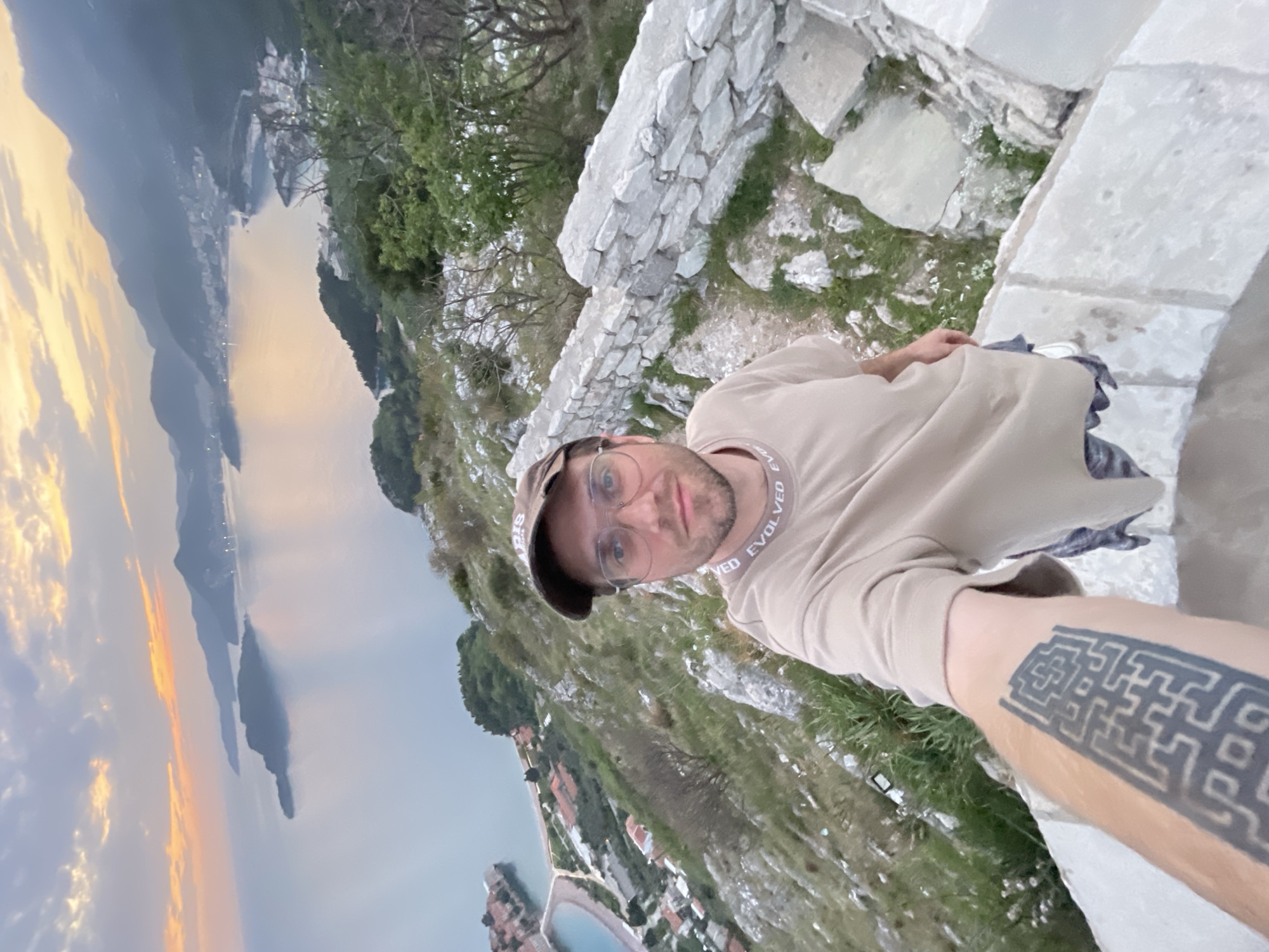 An outdoor selfie captures a man on a stone-walled viewpoint overlooking the coastal landscape of Sveti Stefan, Montenegro, during late afternoon or early evening. The man, seen from the chest up, looks directly at the camera with a neutral expression. He wears a light tan long-sleeved shirt with the word "EVOLVED" visible repeatedly on its collar, a brown baseball cap, and clear-rimmed glasses. A distinctive black, geometric, maze-like tattoo is prominent on his left forearm, extended towards the camera.
Behind him, rugged green hills descend towards a large body of water, likely the Adriatic Sea, reflecting the warm, soft hues of the sky. In the distance, the iconic Sveti Stefan island, densely populated with buildings and connected to the mainland by a narrow causeway, is clearly visible. The sky above is partly cloudy, displaying shades of soft orange and blue, indicative of sunset. The immediate foreground features a rustic stone wall and sparse vegetation.