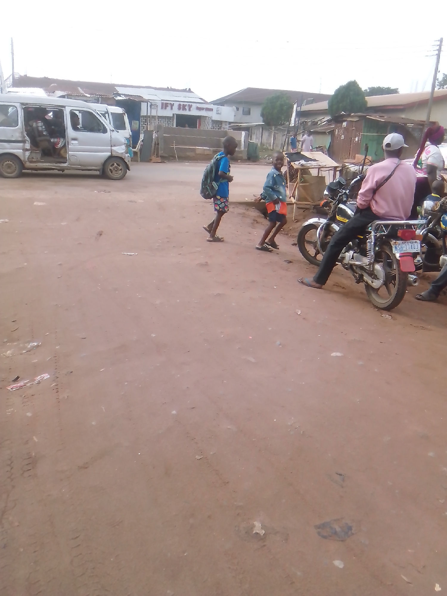 A street scene in an unidentified city. A white van sits to the left. Two children are walking. A man wearing a pink shirt and a baseball cap sits on a motorcycle. Another motorcycle is next to the first. The ground is dirt. There are buildings and other people in the background. A sign that says "IFY SKY" is visible.