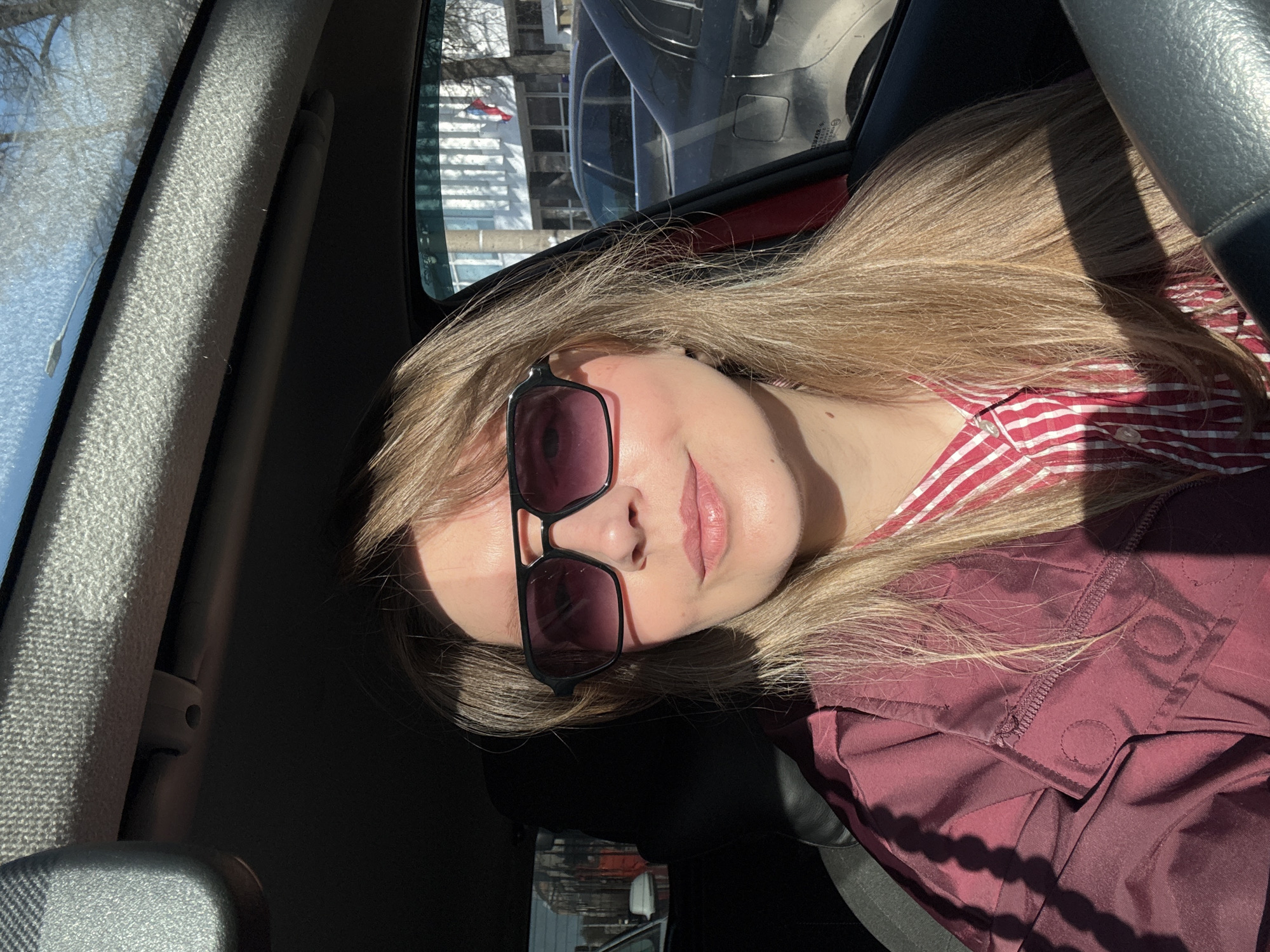 A medium close-up portrait, rotated sideways, captures a young woman sitting inside a car on a sunny day. She has long, straight blonde hair and is wearing dark-framed sunglasses with dark lenses. Her lips are slightly parted, and she has a serene expression.
She is dressed in a red and white striped top, layered under a dark red or maroon quilted jacket. Strong sunlight from the upper left casts distinct shadows across her face, hair, and clothing, as well as parts of the car's interior.
The setting is inside a vehicle. Through the car window on her left, a textured A-pillar and a view of bare tree branches and distant buildings are visible, partially obscured by what appears to be frost or ice on the glass. The window on her right reflects an inverted image of the exterior, showing more buildings and possibly another vehicle. The car's interior features dark surfaces and a visible steering wheel on the right side of the frame.
The image conveys a calm moment during daylight hours, characterized by bright sunshine and potentially cold weather, suggested by the frost and bare trees. This aligns with the context of Izhevsk, Russia, potentially in winter or early spring. No readable text is visible.