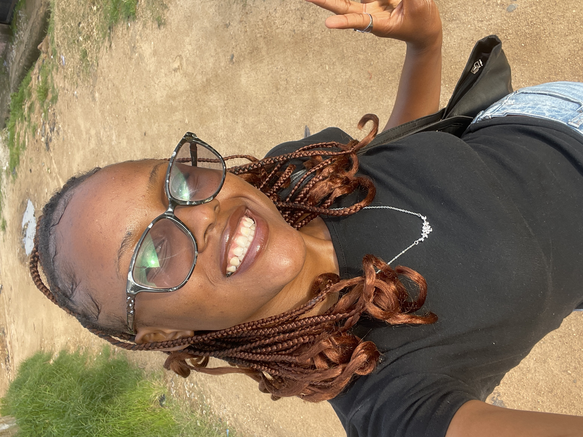 A close-up, high-angle selfie shows a Black woman with braided hair and glasses smiling broadly. She is wearing a black t-shirt and denim jeans, with a black shoulder bag visible. The background consists of a dusty, unpaved ground with patches of dry grass. A hint of greenery is seen in the lower left corner, suggesting an outdoor setting. The lighting indicates it is daytime, likely sunny, given the bright reflections on her glasses and the overall illumination. The woman appears happy and relaxed, capturing a moment of joy. No visible text is present. The context places this scene in Jos, Nigeria.