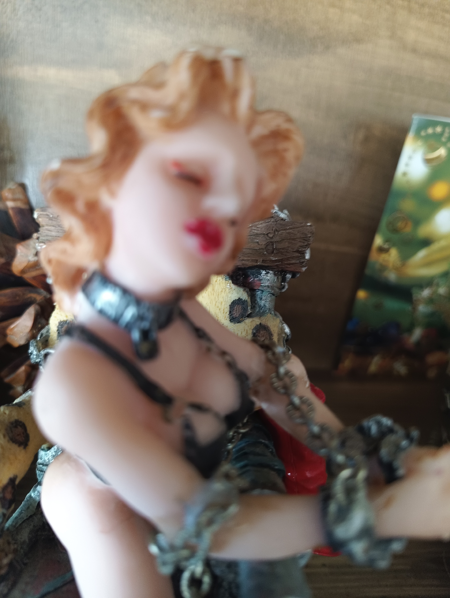 This is a close-up, slightly out-of-focus photograph of a figurine. The figurine appears to be of a woman with blonde, wavy hair, rosy cheeks, and red lips. She is wearing a dark, possibly leather, outfit with chains around her neck and wrists. The figurine is adorned with what looks like a leopard print pattern on its torso. In the background, a wooden surface is visible, along with a blurry green and yellow object that could be a postcard or a picture. The lighting suggests an indoor setting. The overall impression is that of a decorative item, possibly a collectible figurine, placed on a shelf or table.
