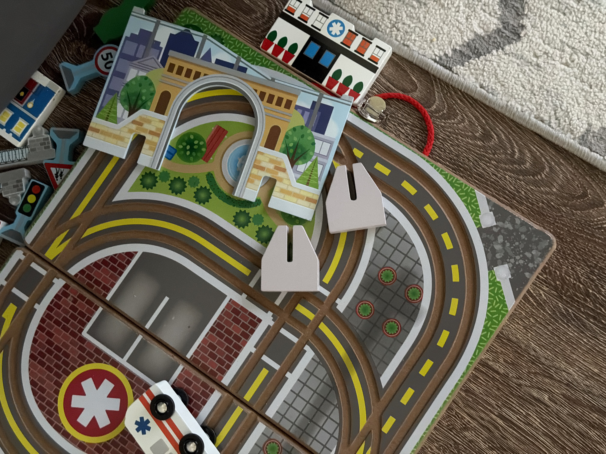 The image displays a wooden toy city play mat on a wooden floor, partially covered by a textured rug. The play mat depicts a city scene with roads, buildings, trees, and traffic signs. A small toy ambulance with red and white stripes and a blue star emblem is positioned on one of the roads, near a building with a red brick facade and a white star symbol within a red circle. Several other toy elements are scattered around, including a small toy house, traffic lights, and road signs. A red string is attached to one of the toy buildings. The scene is lit from above, casting subtle shadows. There are no people visible in the image. The location in the city of Katy, United States is not discernible from the image.