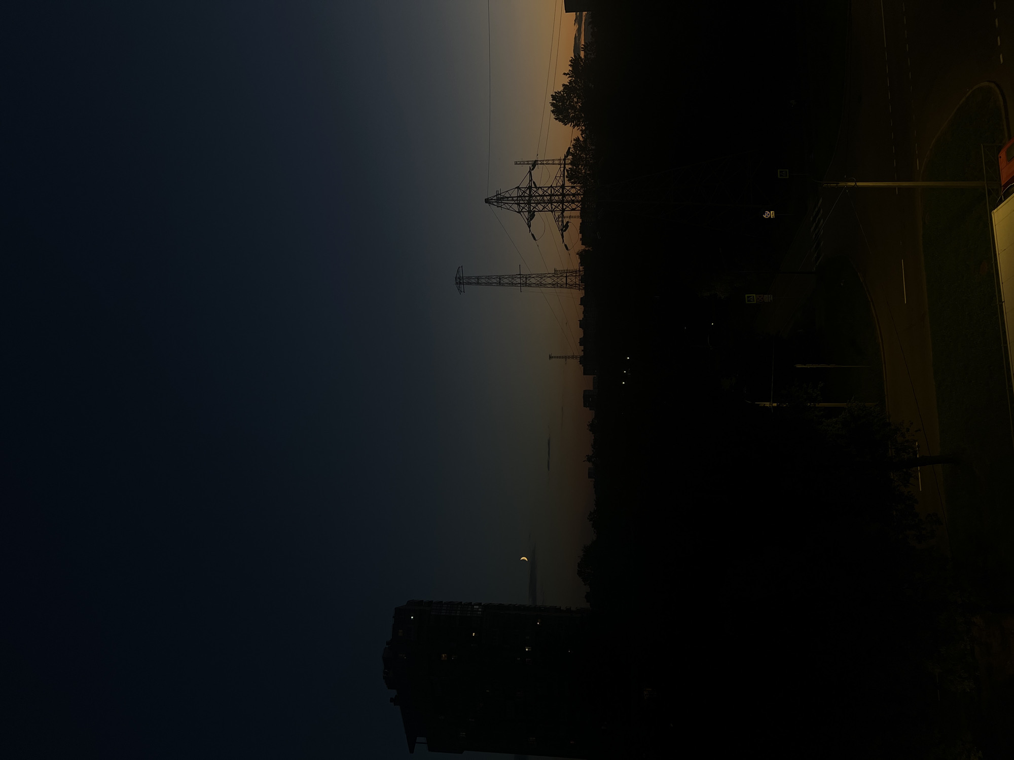 The image depicts a silhouetted city skyline at dusk in Sinyavino, Russia. A tall, dark building is visible on the left, with a few illuminated windows. To its right, a thin crescent moon hangs in the darkening sky. In the middle ground, the silhouettes of trees and buildings form a dark band across the horizon. Prominently featured are several high-voltage electricity pylons, with one particularly large and complex structure dominating the center-right of the frame. The sky transitions from a deep blue at the top to a faint orange and yellow glow near the horizon. The foreground is almost entirely dark, with only a hint of a road and some faint lights, suggesting it is night or very late dusk. There are no visible people or animals in the frame.
