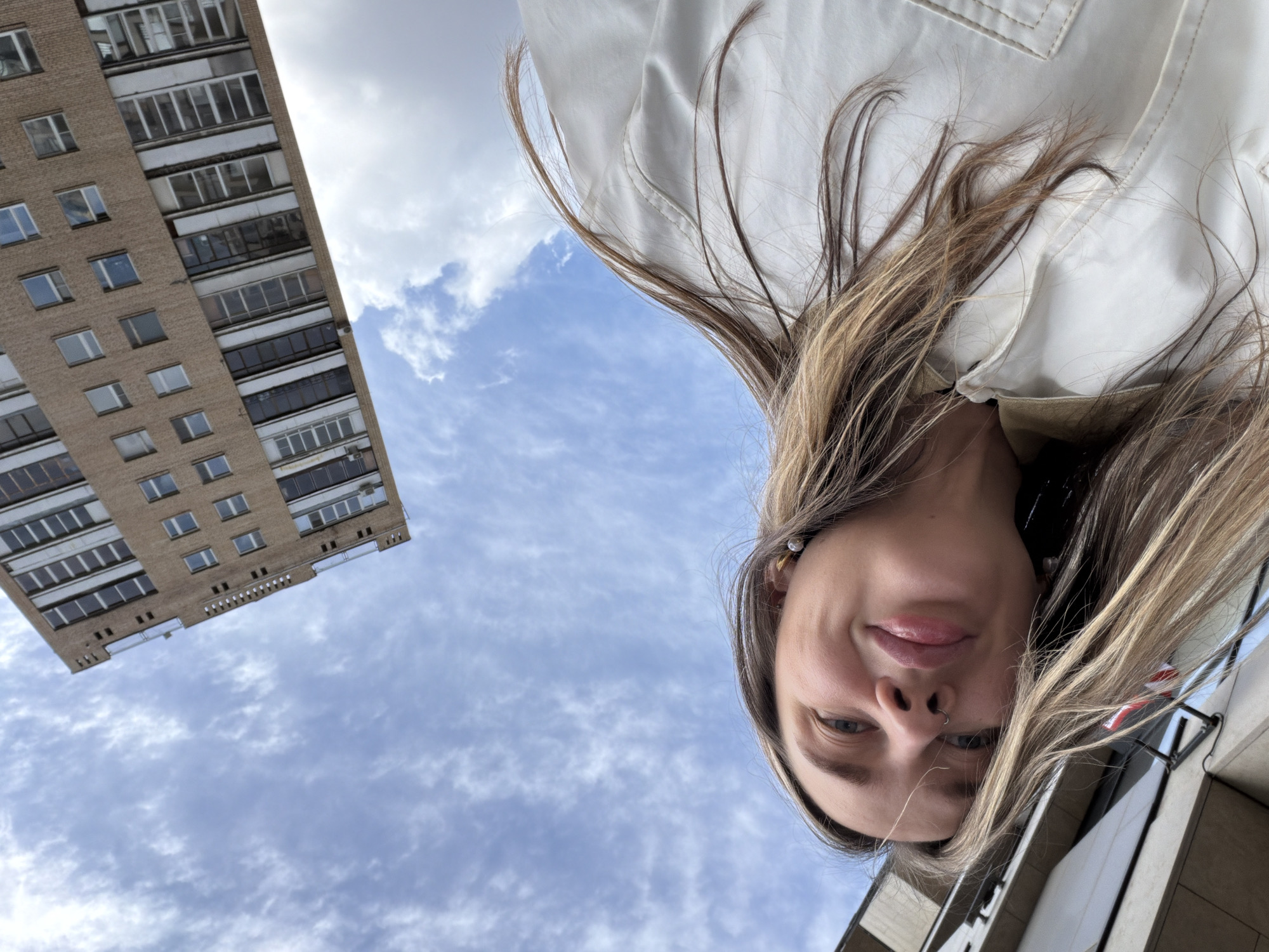 A low-angle selfie, taken outdoors in Moscow, Russia, features a woman with light brown and blonde hair, wearing a white jacket. The sky is a clear blue with light, patchy clouds. In the upper left corner, a brick apartment building with numerous windows is visible. The woman's face is the most prominent subject, with a partial view of another building at the bottom right.