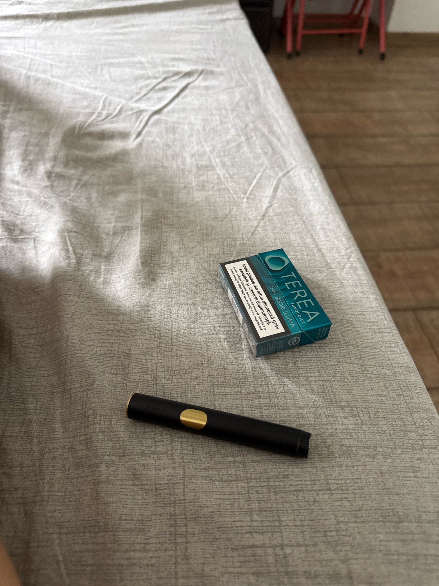 The image displays a close-up, high-angle view of a dark, cylindrical electronic smoking device with a gold accent, resting on a textured, light-colored fabric surface, possibly a bedsheet or tablecloth. To the right of the device, a small, rectangular pack of "TEREA TURQUOISE" tobacco sticks for "IQOS ILUMA" is positioned at an angle. The pack features teal and white branding and warning text in Romanian. The background shows a blurry glimpse of a wooden floor and what appears to be a pink chair, suggesting an indoor setting. There are no people visible in the image. The scene is static and does not suggest any specific location in Chișinău, Moldova, beyond being an indoor space.