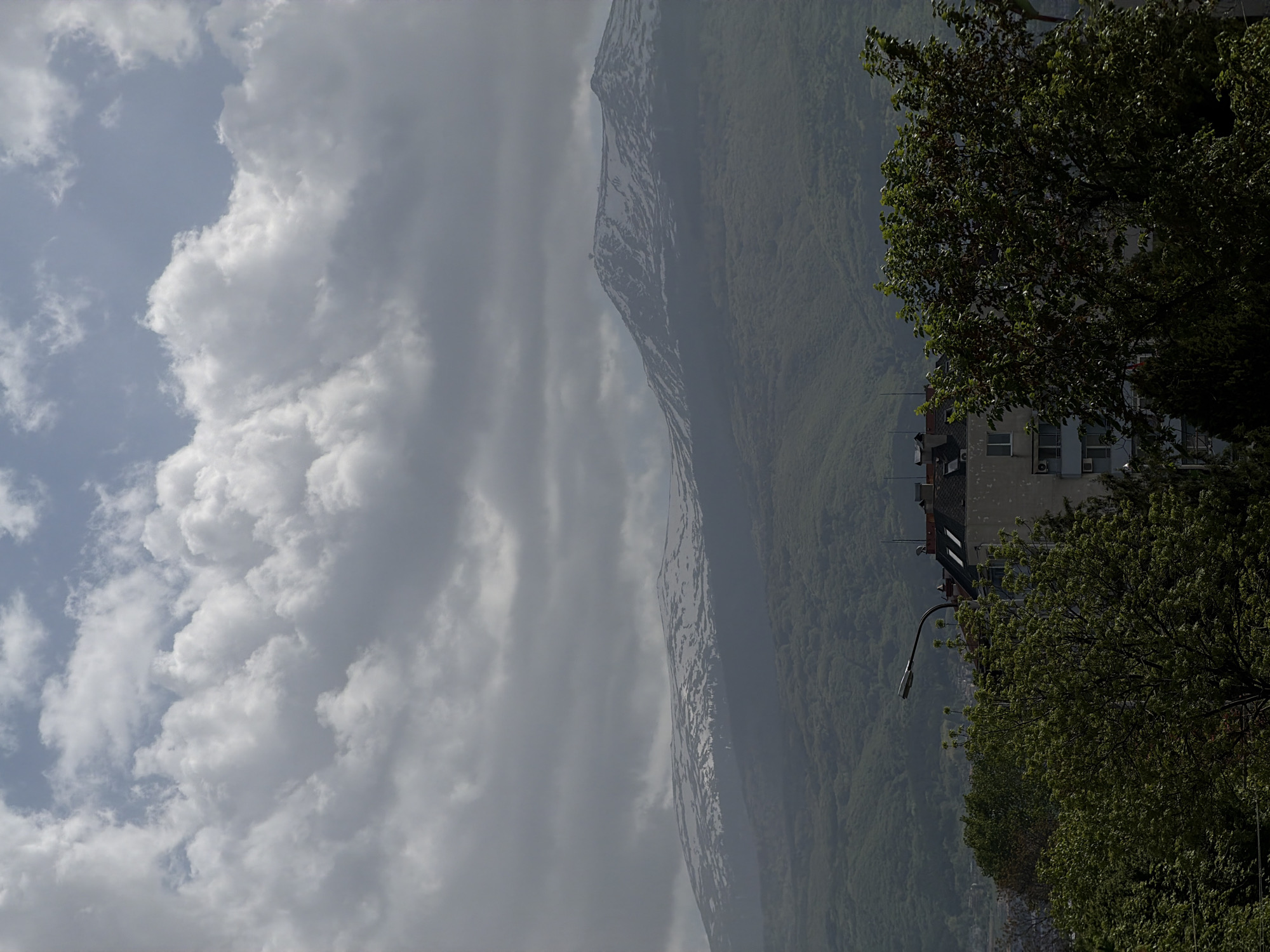 The image shows a landscape with a mountain, sky, clouds, trees, and a building. The mountain is partially covered in snow and appears to be adjacent to a city, which is only partially visible. A street lamp is visible near the building. The sky is filled with clouds, ranging from light to dark grey. The trees are full of green foliage. The location is possibly a city with a mountain backdrop.