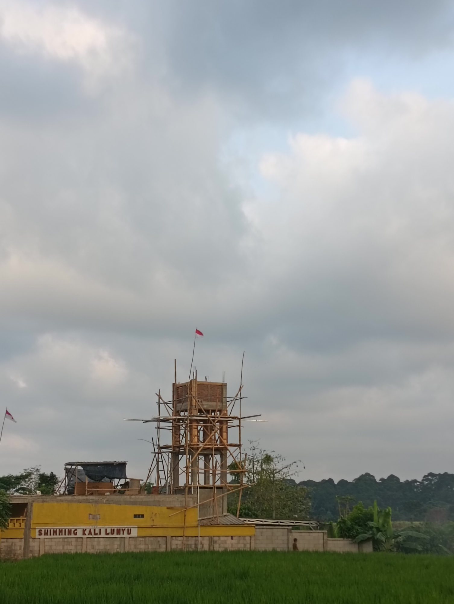 This image depicts a water tower under construction in Tayu, Indonesia. The unfinished tower, made of wood scaffolding and brickwork, is the central subject, topped with a small red and white flag. To the left of the tower is a yellow building with signage that reads "SWIMMING KALI LUNYU". The building appears to be a commercial establishment, possibly a swimming facility, given the signage. In the foreground, a lush green field of rice paddies stretches across the bottom of the frame. The sky is overcast with scattered clouds. A lone figure can be seen near the yellow building, walking in front of a grey concrete wall. The background is dominated by a dense line of trees, suggesting a rural or semi-rural setting.
