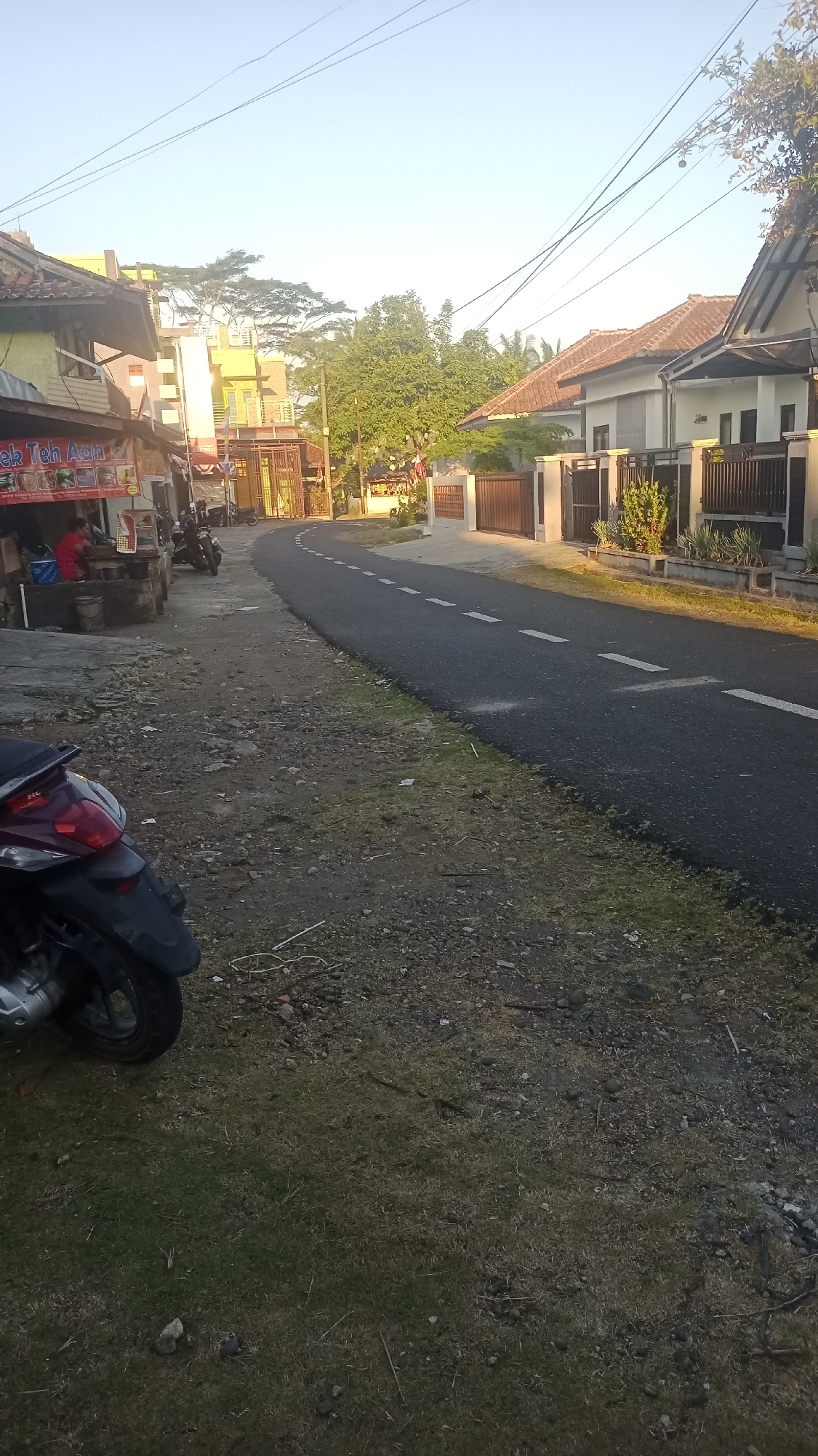 The image depicts a street scene in Pakemitan, Indonesia, bathed in the soft light of either early morning or late afternoon. A paved road curves to the right, bordered by a grassy, dirt shoulder. On the left side of the road, a small food stall with a red awning and seating area is visible, with a person seated at the stall. Several motorcycles are parked nearby. Beyond the stall, a multi-story building with yellow and green accents stands, alongside a tall, gated entrance. Across the street, on the right, are modern residential houses with tiled roofs and fences. Lush green trees are scattered throughout the landscape, and utility wires crisscross the clear, pale sky. The foreground of the image is dominated by the rear wheel and fender of a parked black motorcycle.