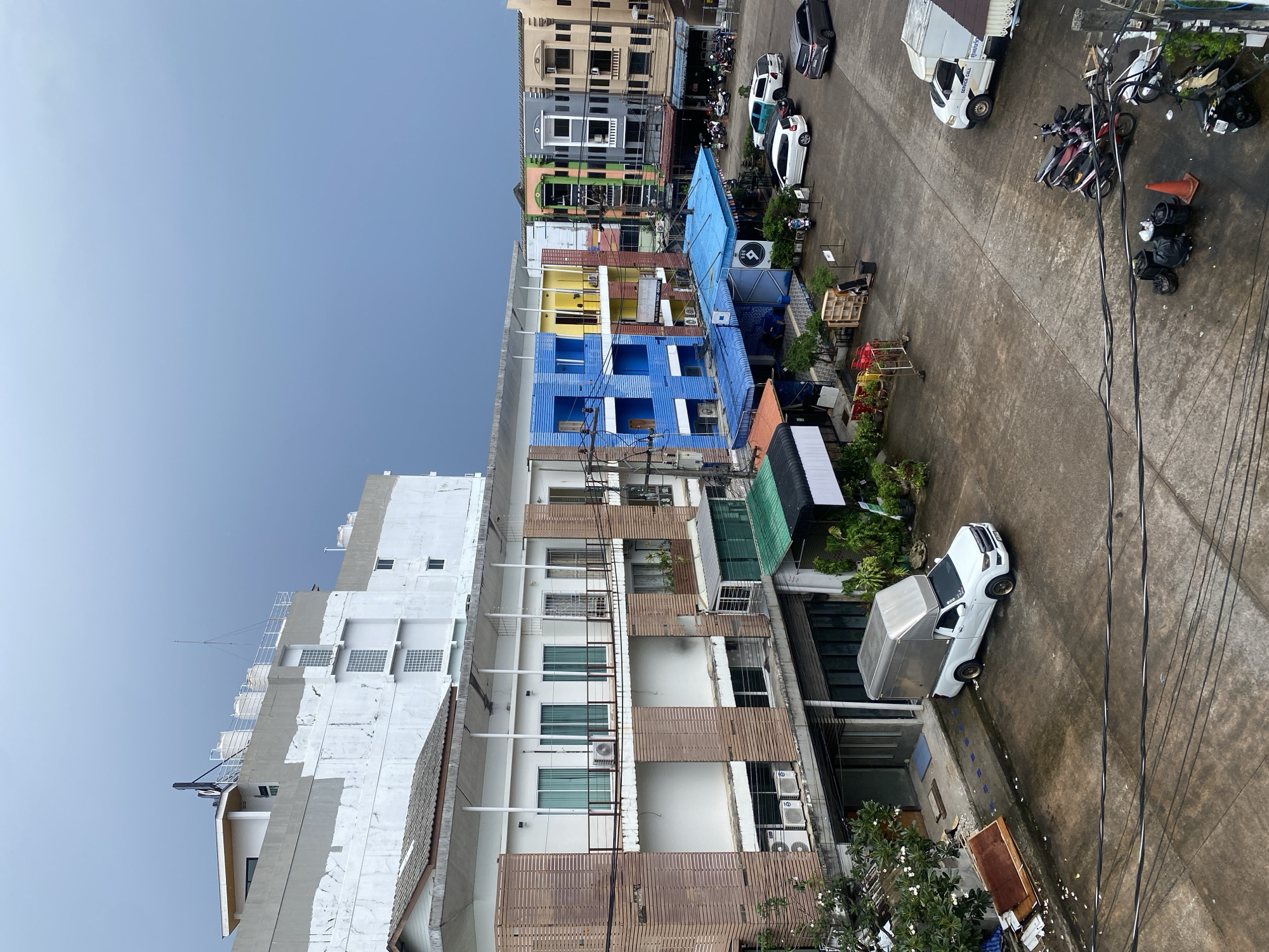 An exterior, high-angle, medium shot captures a cloudy day in Phuket City Municipality, Thailand. The scene depicts a courtyard in front of a multi-story building with a light gray and white facade. A white pickup truck with a silver cargo box is parked in the courtyard, alongside several motorcycles and other vehicles. The courtyard is paved with concrete and appears wet, suggesting recent rain. Buildings of various colors and architectural styles line the courtyard, including a bright blue building with yellow accents. Some of the buildings have awnings and balconies, and there are a few trees and potted plants scattered around. Electrical wires are visible overhead, crisscrossing the sky. The overall impression is of a somewhat utilitarian urban environment, possibly a commercial or residential area.