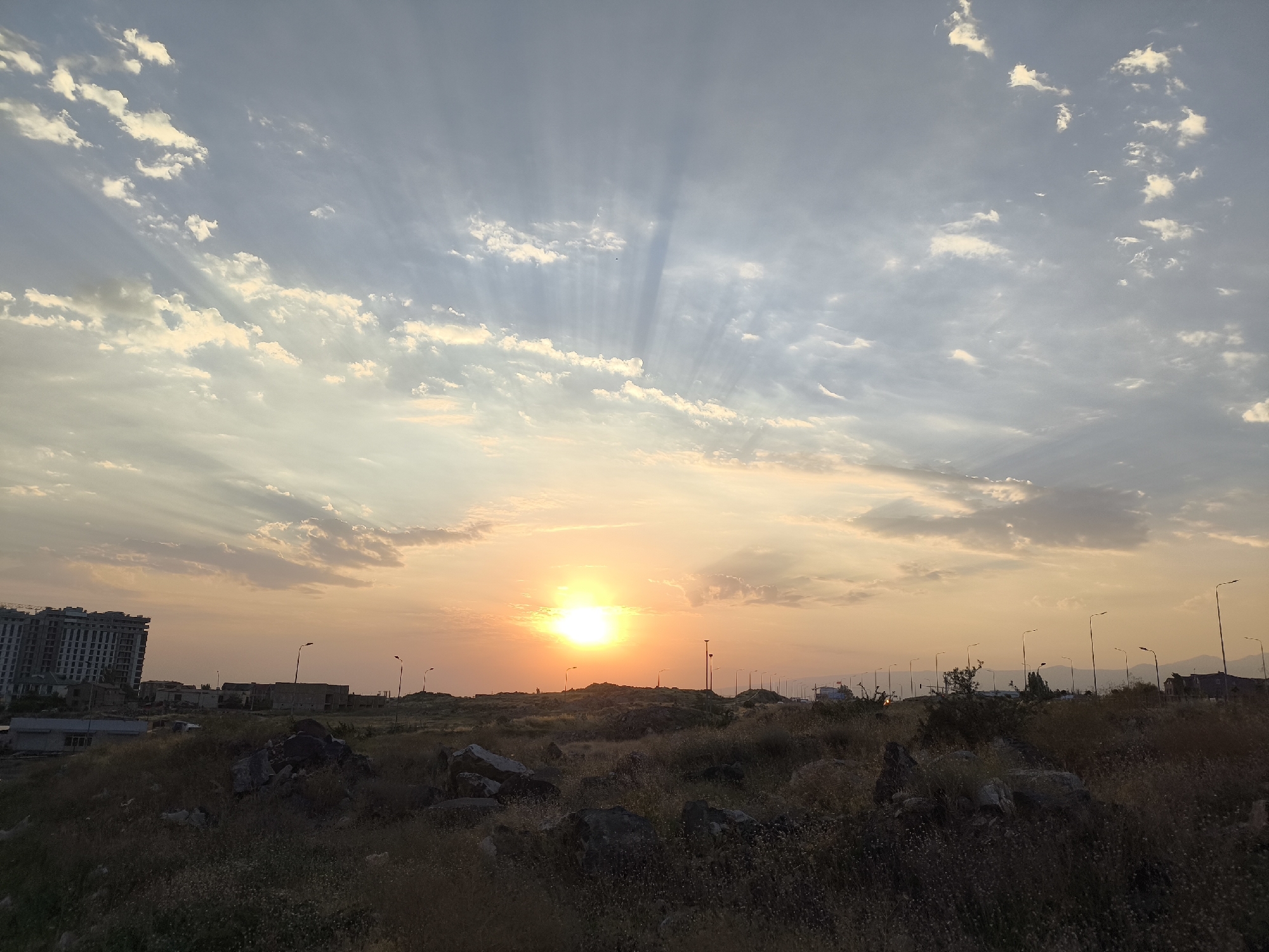 The image captures a sunset over a semi-urban landscape in Yerevan, Armenia. The sun, a bright orb, is partially obscured by wispy clouds, casting dramatic crepuscular rays across the sky. The foreground is dominated by dry, rocky terrain with sparse vegetation. In the middle ground, a construction site with a partially built, multi-story building is visible on the left, along with several low-rise structures and streetlights. A road with some vehicles can be seen in the distance, suggesting proximity to an urban area. The sky transitions from a pale blue at the top to hues of orange and yellow near the horizon, indicating the sun's low position.