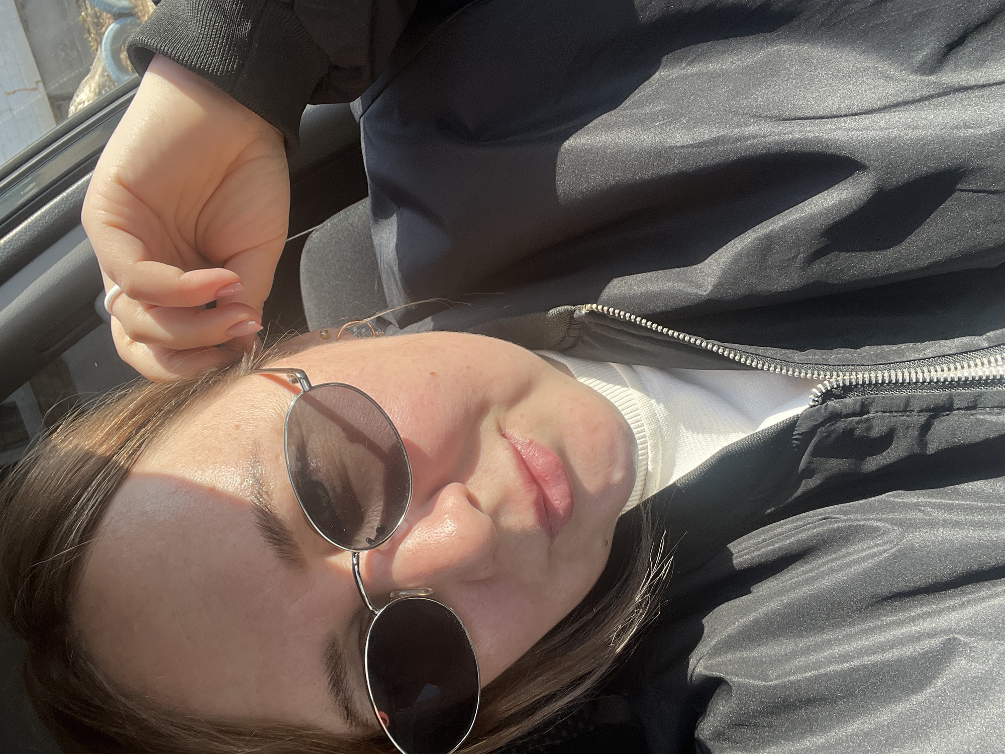 A person is lying down outdoors, possibly in a car, in Chaikovsky, Russia. The person is wearing sunglasses, a white turtleneck sweater, and a black jacket. Their left hand is raised. The sun is shining.