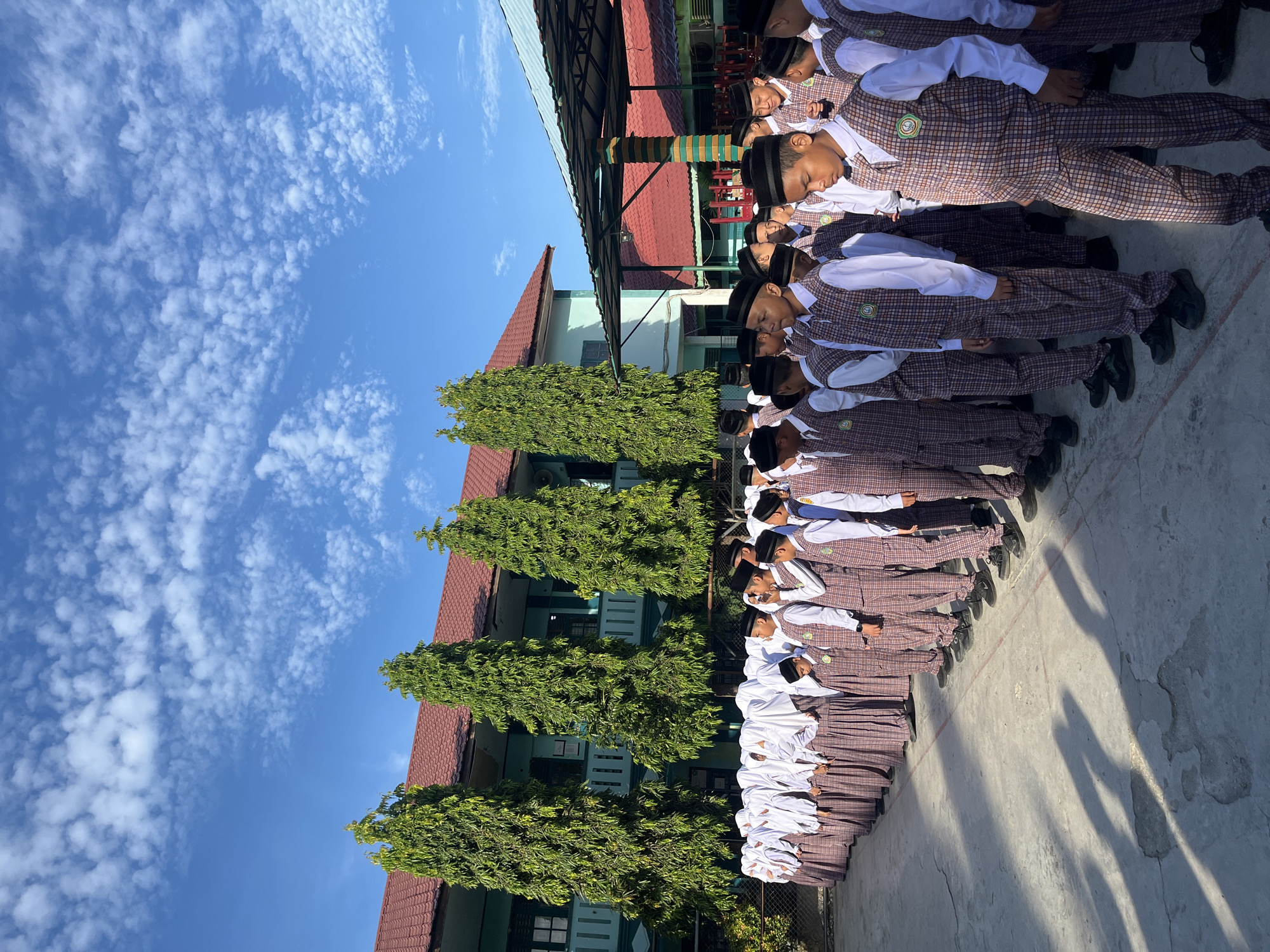 A group of male students, dressed in white shirts, plaid vests, plaid pants, and black skullcaps, stand in formation on a paved outdoor area. They are in a schoolyard, with a light blue building with a red roof and tall green trees visible in the background. A covered walkway with a red roof is also present. The sky is clear blue with scattered white clouds. The students appear to be in Lhokseumawe, Indonesia.