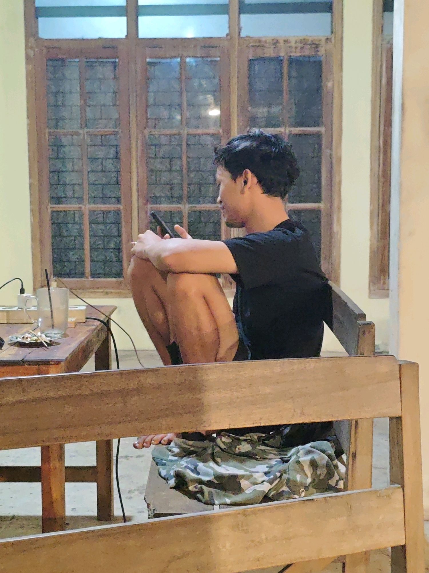 The image depicts a young man sitting on a wooden chair in front of a wooden table, indoors. He is wearing a black t-shirt and camouflage shorts. He is holding a smartphone in his hands and appears to be looking at it. On the table, there is a glass of water with a straw, a small ashtray with cigarette butts, and some other items. Behind the man is a wooden window with multiple panes. The environment suggests a relaxed setting, possibly a cafe or a home. The overall lighting is dim, indicating it might be evening or an indoor space with limited natural light. The location is specified as Ngasem, Indonesia.