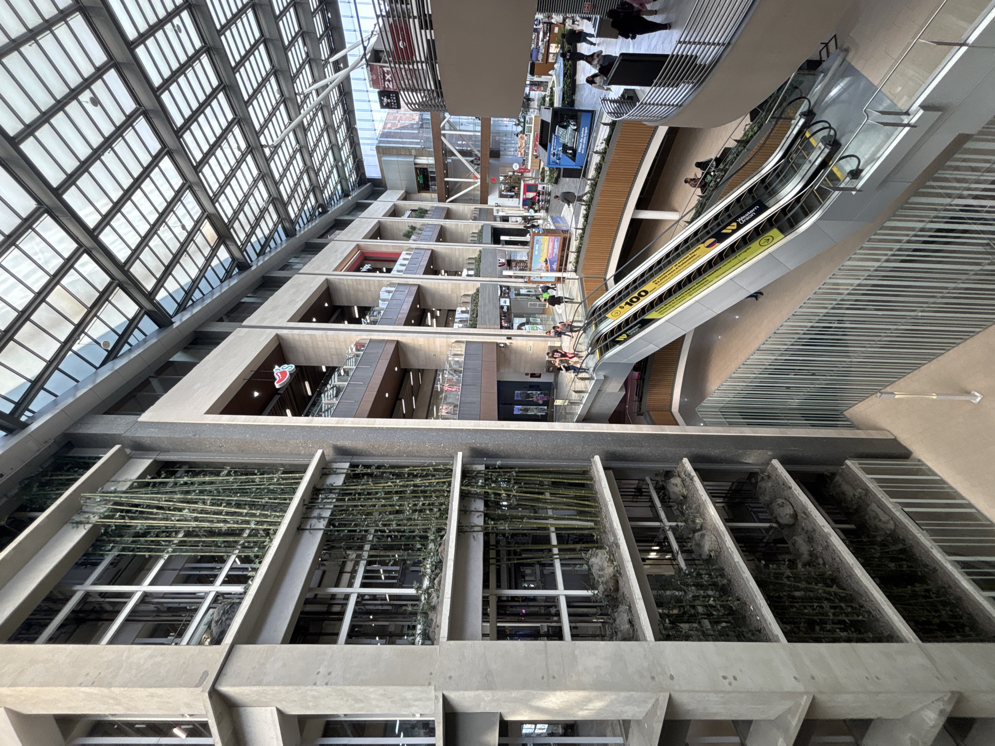 The image depicts the interior of a multi-story shopping mall in Mexico City, Mexico. The building features a slanted glass ceiling, allowing natural light to illuminate the space. There are multiple levels, with various stores visible. Escalators provide access between floors. Several people are visible walking, shopping, and riding the escalators. Concrete architectural elements, including windowed ledges, create a patterned façade. A green plant arrangement adorns the lower section of the building.