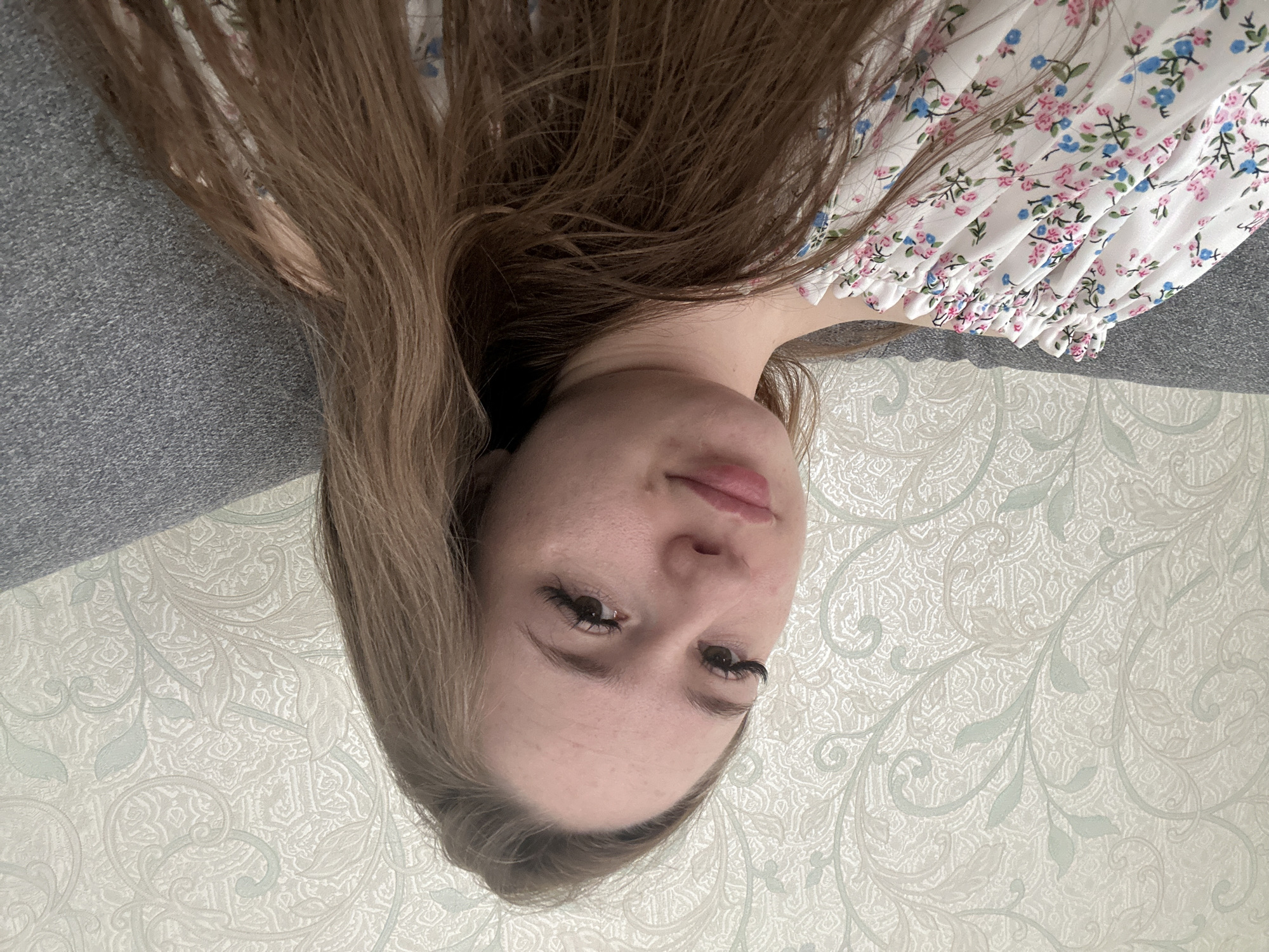 An upside-down, overhead shot depicts a young woman lying down, possibly on a bed or sofa. Her head and upper torso are visible, with her long, light brown hair fanning out around her. She has a calm, neutral expression, with dark eyes accentuated by long, dark eyelashes. Her lips are slightly parted, revealing their natural pink color. She is wearing a white garment adorned with a delicate floral pattern of small pink and blue flowers, featuring gathered fabric around what appears to be a neckline or shoulder.
To the upper left of the frame (if viewed upright), a textured gray surface, likely upholstery, is visible. The lower right portion of the image shows a wall covered in light-colored wallpaper. This wallpaper features an elaborate, ornate pattern of scrolls, leaves, and abstract designs in pale green and cream hues. The lighting is soft and even, suggesting an indoor environment during the daytime. No text is visible in the image.