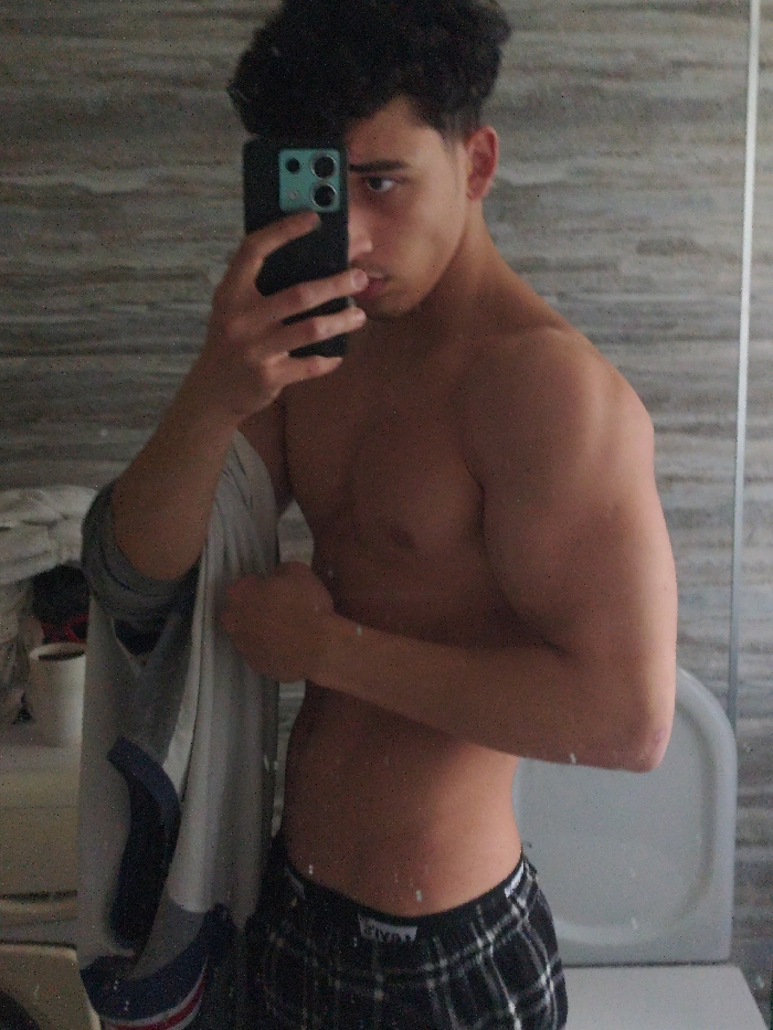A male individual is captured in a mirror selfie, positioned in what appears to be a bathroom. He has dark, curly hair and an athletic build. His upper torso is bare, displaying developed musculature. He wears black and white plaid boxer shorts; a brand logo is visible on the waistband. His right hand holds a dark-cased smartphone, used to capture the mirror reflection and partially obscuring the right side of his face. His left arm is flexed, hand clenched into a fist, displaying the bicep. A light-colored garment, featuring white, grey, and blue sections, is draped over his left forearm. The background, a mirror reflection, shows light grey, horizontally laid wall tiles. A white toilet fixture is visible on the right side of the reflection. A white cylindrical container is partially visible on a surface to the left. The precise location within As-Salihiya Municipality, Syria, is not discernible from the visual information provided.