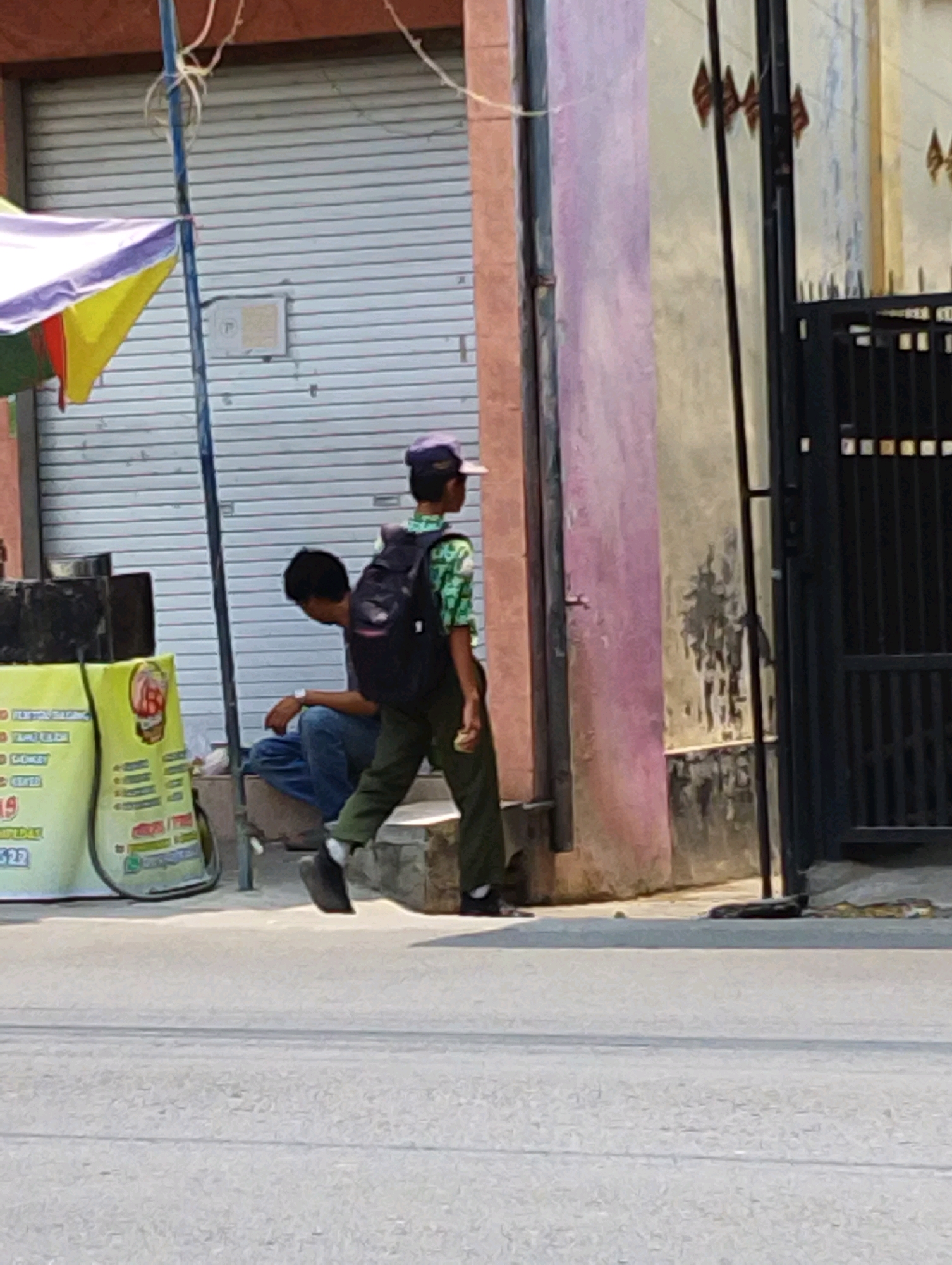 In Gempeng, Indonesia, on a bright day, two young males are observed in front of a building with a grey roll-up door and a pink and beige facade. A tall, thin blue pole, likely a street sign or utility pole, stands to the left of the pink section of the building, supporting a colorful awning with yellow, purple, and red fabric sections. The younger male on the right is walking away from the camera, wearing a green and white patterned shirt, dark green pants, and a dark backpack. He has a purple and light pink cap on his head. The second male is sitting on a concrete ledge to the left, facing away from the camera, wearing blue jeans and a dark shirt. He has a watch on his left wrist. To the far left of the image, a yellow sign with red and black lettering is partially visible, possibly a business or information board. The ground is asphalt. A black metal gate is visible on the far right of the image.