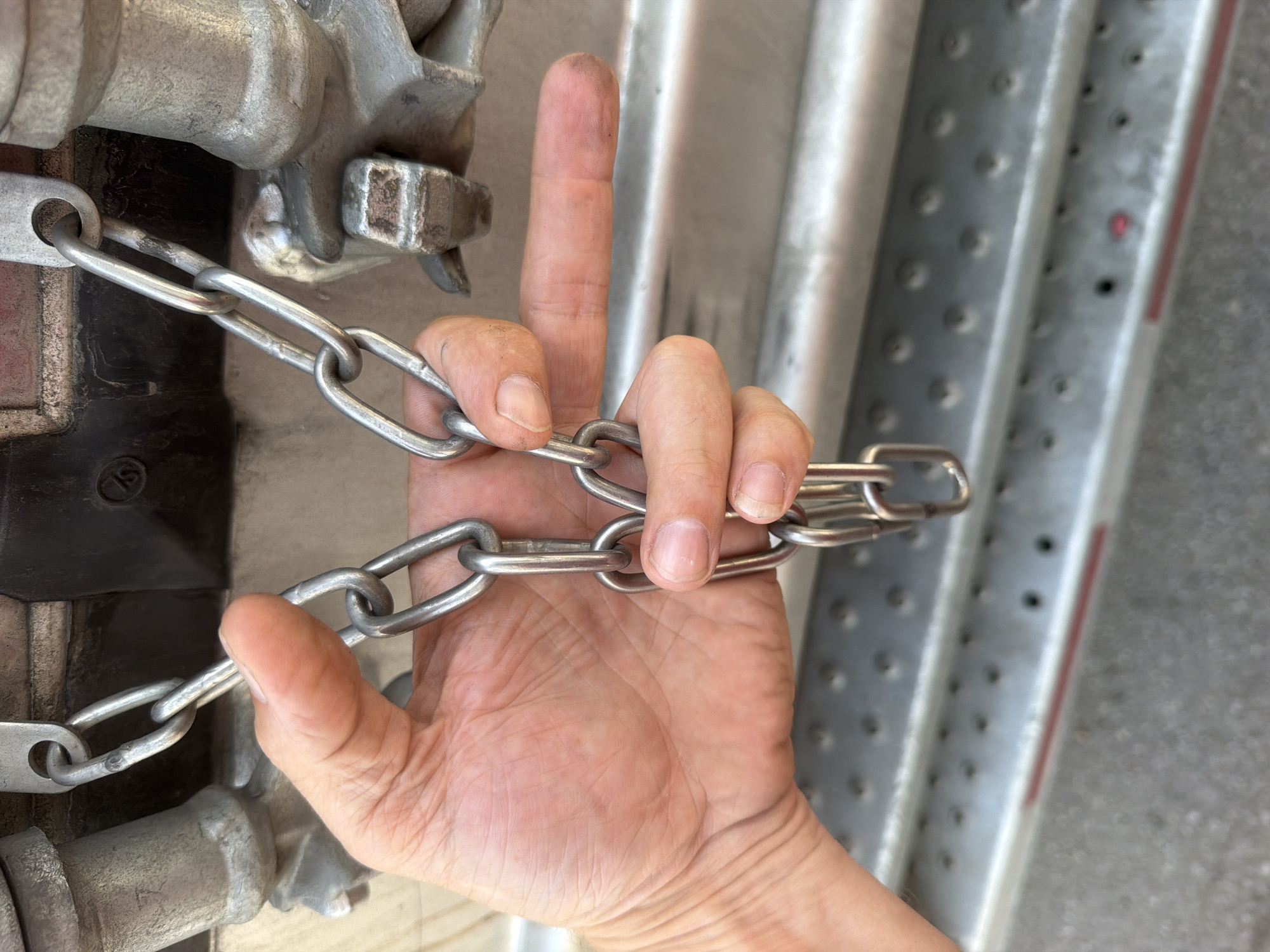 A person's hand and fingers are shown in a close-up shot, gripping a metal chain. The chain is attached to a larger metal object in the background, which appears to be part of a vehicle or industrial equipment. The background also shows a textured metal surface with numerous circular indentations, suggestive of a ramp or a walkway. The ground beneath is visible and appears to be asphalt. The lighting suggests it is daytime. No specific location in the city of Lebanon, United States of America, can be identified from the visual information provided.