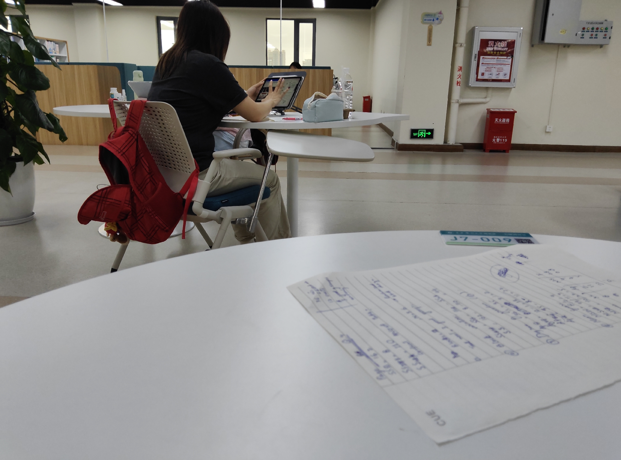 This media file shows an indoor scene, likely a library or study area, with several tables and chairs. A woman is seated at a table, facing away from the camera, and appears to be using a tablet. She is wearing a black t-shirt and light-colored pants. A red jacket with a plaid pattern is draped over the back of her chair. To her right, on the table, is a light blue pouch and a bottle of water.
Another table in the foreground is mostly out of focus, but a sheet of lined paper with handwritten notes and calculations in blue ink is clearly visible on it. A small green and white sticker with the text "J7-009" is also on this table.
In the background, there are bookshelves and partitions. On the wall, there is a red fire extinguisher box with Chinese text that includes "灭火器箱" and "火警119," indicating it's a fire extinguisher cabinet and emergency number. There is also a sign with Chinese characters, "消火栓," which means "fire hydrant." An exit sign with an arrow is visible on the wall to the left. The lighting suggests it is daytime, but there are no strong indicators of the time of day or weather outside. The overall atmosphere appears quiet and focused, consistent with a study environment.