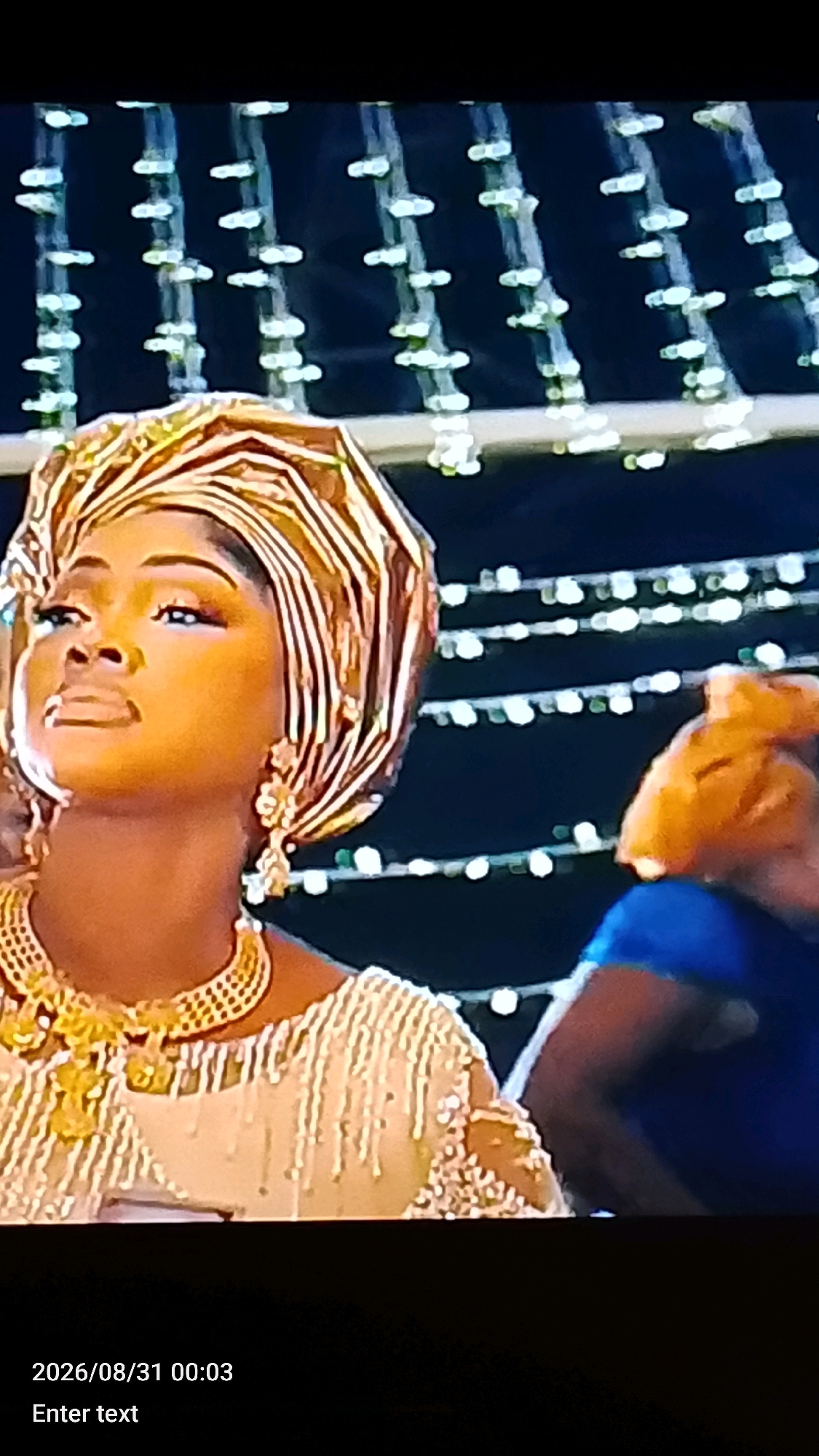 The image shows a close-up, medium shot of a woman from the shoulders up, facing left and looking upwards. She is wearing a golden headwrap and elaborate gold jewelry, including a necklace and earrings. Her dress is intricately beaded and white or cream-colored. Behind her, out of focus, is another person wearing a blue garment, and in the background, there are strings of white lights against a dark backdrop. The lighting is dim, creating a festive or ceremonial atmosphere. The timestamp at the bottom of the image reads "2026/08/31 00:03". This image does not contain any specific visual cues that would allow for its location to be identified in Lokoja, Nigeria.