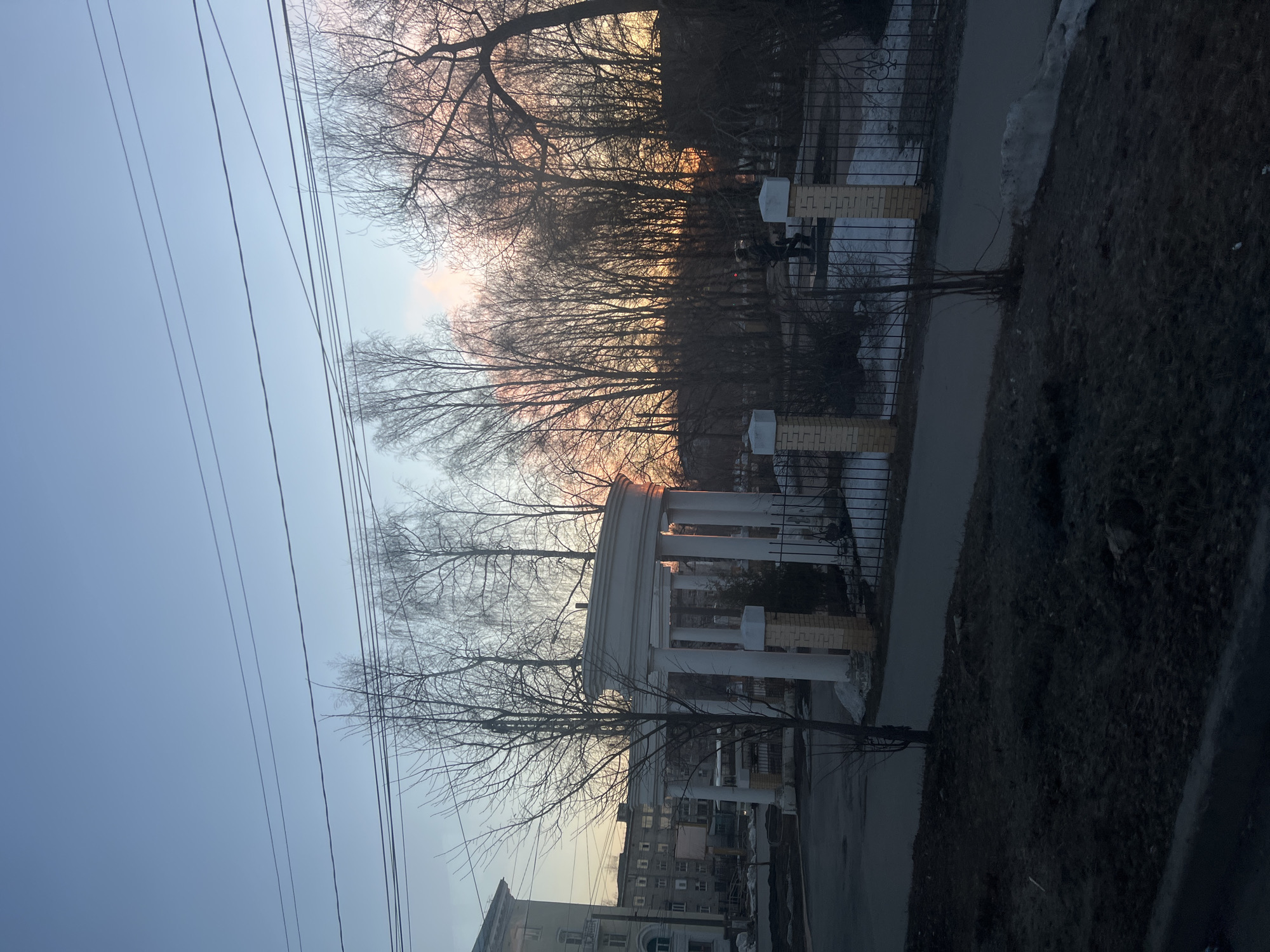 The image depicts a scene at dusk or dawn in Usolye, Russia, featuring a classical rotunda with white columns and a dark-colored roof. The rotunda is positioned along a paved path, which is bordered by a dark, possibly muddy, area and a metal fence. Beyond the fence are bare trees silhouetted against a sky with faint orange and pink hues, indicating sunrise or sunset. In the foreground, utility wires stretch across the sky. Low buildings are partially visible in the lower portion of the frame. No people are visible in the scene.