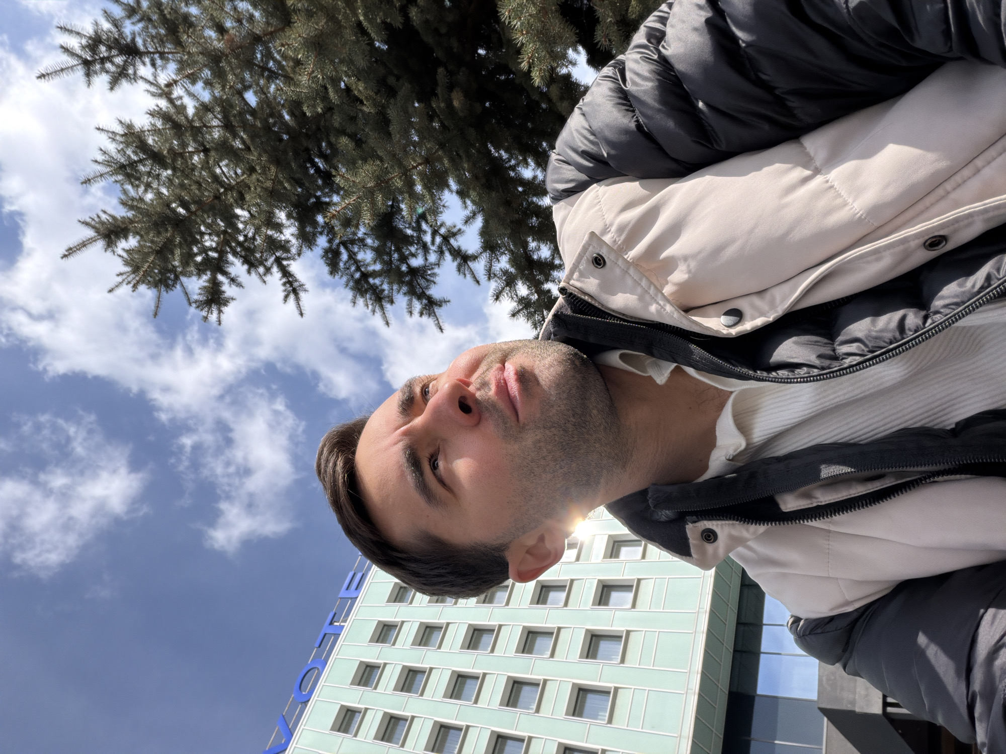 An outdoor, low-angle shot, rotated 90 degrees clockwise, features an adult man looking upwards with a neutral expression. His head is positioned on the right side of the frame. He has short dark hair and noticeable stubble. He is dressed in a two-toned puffer jacket, which is black on the upper half and a light beige or off-white on the lower half, worn over a white ribbed top. Bright sunlight highlights his face, creating a sun flare near his right ear.
The background consists of a clear blue sky with scattered white clouds. To the upper left, dense, dark green branches of a coniferous tree are visible. To the lower right, a modern building of light teal or mint green color, featuring numerous rectangular windows, rises vertically. Partially visible blue lettering on the building spells "OTE," likely part of a word like "HOTEL." The scene suggests a sunny daytime setting in an urban environment.