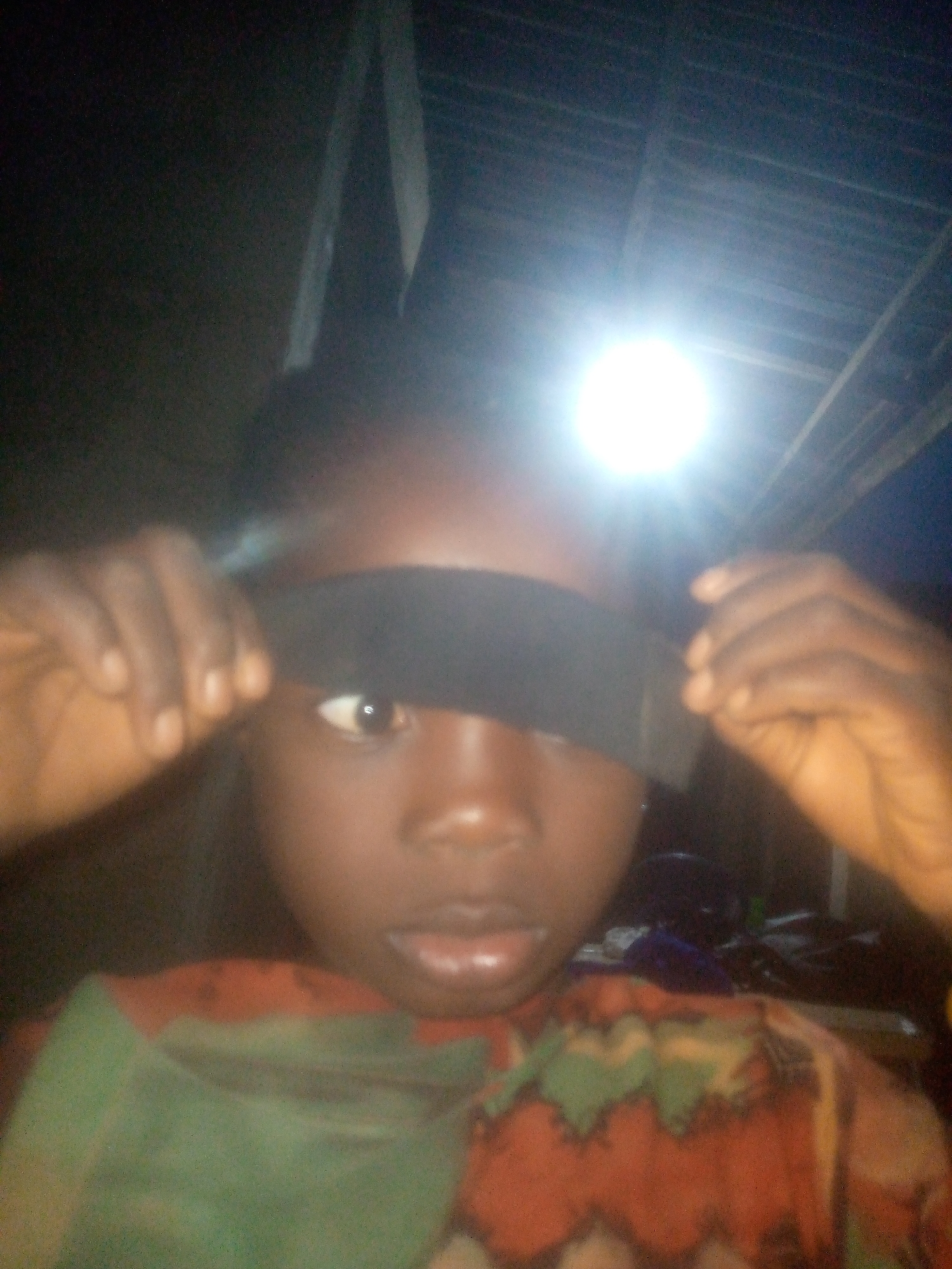A dimly lit, close-up portrait captures a young person, likely a child, with dark skin, looking directly at the camera. The subject is holding a dark, horizontal band of fabric or material across their face with both hands, obscuring one eye and much of the forehead. Only one eye, with a visible pupil and iris, can be seen peering from beneath the band. The mouth is slightly open.
A very bright, circular light source, likely a camera flash or a powerful overhead lamp, dominates the upper part of the image, casting intense illumination downwards. This harsh artificial light suggests the photograph was taken in a very dark environment, possibly indoors at night. The background is largely obscured by shadow, but faint outlines of what appear to be ceiling structures or beams are visible in the top corners. The subject is wearing patterned clothing featuring green, orange, and possibly brown hues in an abstract or geometric design. No text is visible in the image.