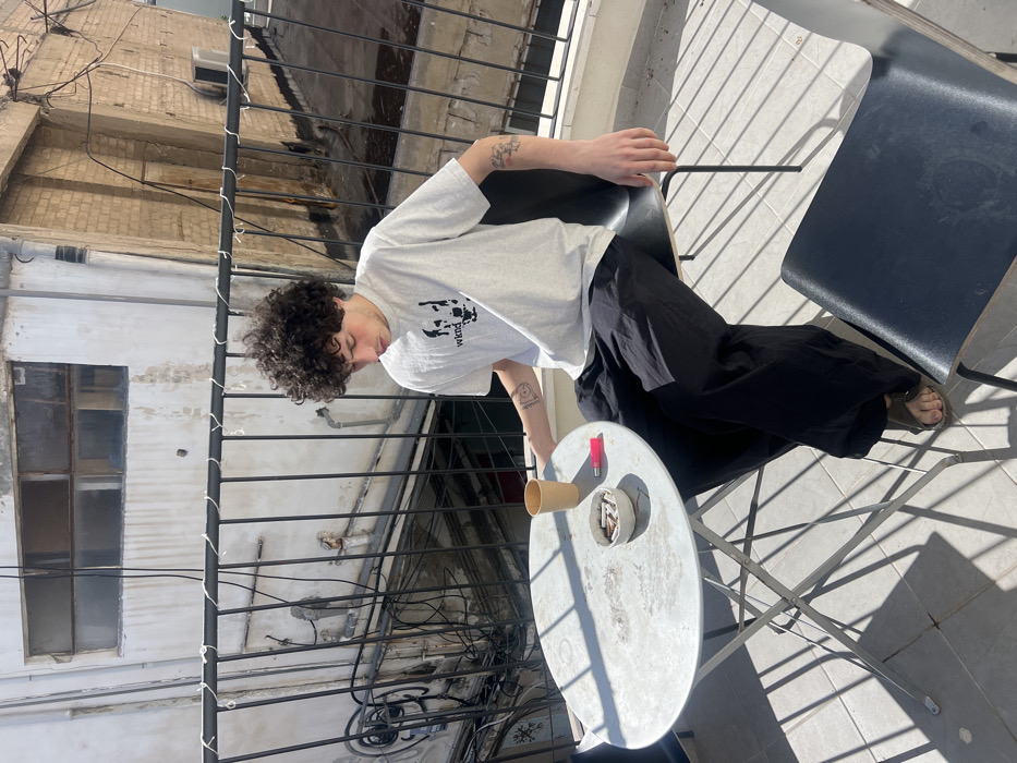 The image depicts a young adult male seated on a tiled balcony in an urban setting, identified as Tel-Aviv, Israel. The male has dark, curly hair and is wearing a light grey t-shirt with a black graphic and "wknd" text, dark loose-fitting pants, and dark sandals. Tattoos are visible on both forearms. He is positioned leaning forward, looking down towards a small, round, light-colored metal folding table.
On the table, there is a single light brown disposable cup, a small light-colored ashtray containing several cigarette butts, and a pink disposable lighter. The male's left hand is near the cup, and his right hand rests on his lap or thigh. He is seated on a dark-colored, possibly wooden and metal chair; a second identical chair is partially visible in the foreground to the right, unoccupied.
The balcony features a black metal railing with vertical bars and horizontal supports. Behind the railing, an intricate network of electrical wires and pipes is visible against the weathered exterior of adjacent buildings. The background features several multi-story buildings; one directly behind is light-colored with multiple obscured windows and visible conduits, while another to the right displays reddish-brown brickwork and an external air conditioning unit. The scene is brightly lit by natural sunlight, casting sharp shadows from the railing and furniture onto the tiled floor.