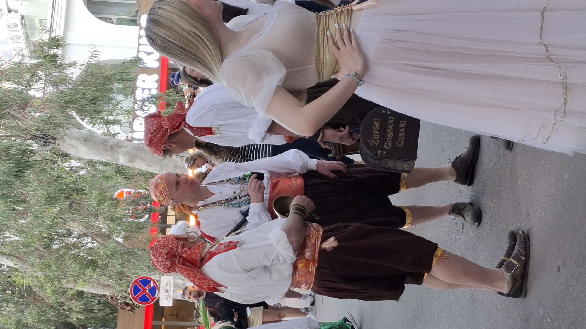 In Budva, Montenegro, a group of individuals is dressed in traditional attire. Three men are wearing red headscarves, white shirts, brown pants, and brown shoes. One of the men is holding a brass bell. A woman in a white dress stands to the side. A sign with a red circle and blue "X" is visible. Another sign reads "Zupski Gosari Gusari." A tree stands in the background, and a building with orange lettering can be partially seen.