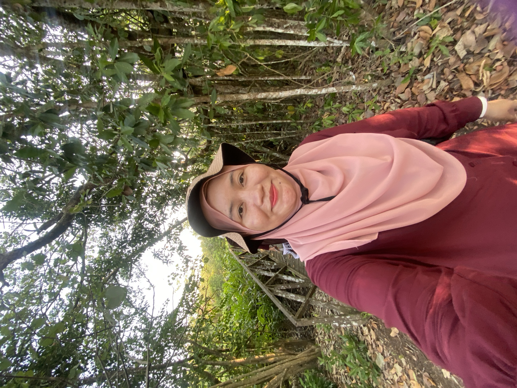 In a sun-dappled forest setting, a woman wearing a maroon long-sleeved shirt, a light pink hijab, and a wide-brimmed hat takes a selfie. She is positioned in the foreground, slightly off-center to the right. Behind her, a wooden walkway with railings meanders through lush green vegetation and trees with textured bark. The ground is covered in fallen leaves and some scattered greenery. The sunlight filters through the dense canopy, creating a bright, natural ambiance. The location is identified as the city of Sungai Pitung, Indonesia.