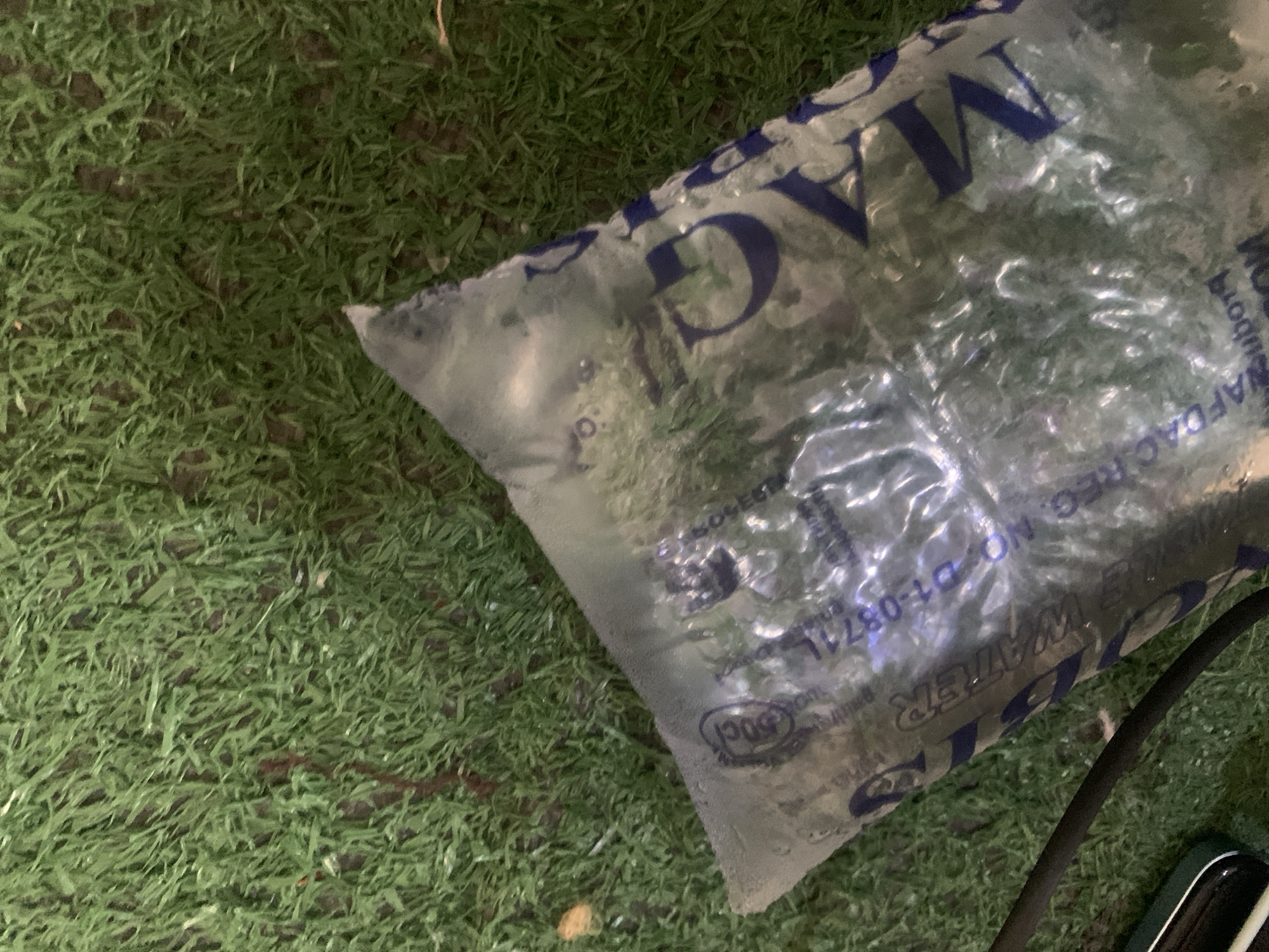 The image shows a clear plastic bag filled with water, lying on a patch of artificial turf. The bag has blue lettering on it, with some text visible. The words "MAG" and "WATER" are clearly legible. Part of a registration number, "D1-0871L", and a seal with "DOC" inside it are also visible. The bag appears to be slightly frosted, suggesting it might be cold. The artificial turf is a vibrant green, with some brown earth showing through the blades. The lighting in the image is somewhat dim, casting shadows and obscuring some details. The overall scene suggests a casual or outdoor setting. There are no people or animals visible in the image. The focus is on the object itself and its immediate surroundings.