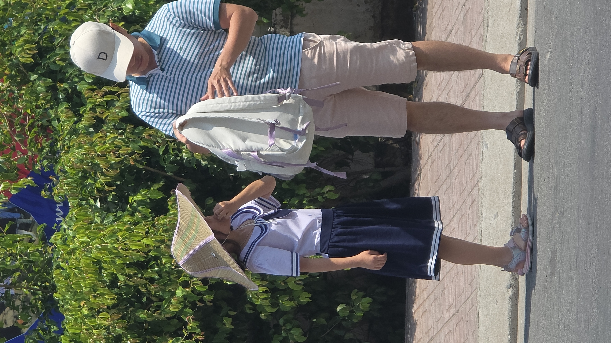 The image shows an adult male and a young girl standing in front of a lush green plant in Nha Trang, Vietnam. The adult is wearing a white baseball cap with a "D" on it, a blue and white striped polo shirt, tan shorts, and black sandals. He is looking into a white backpack. The young girl is wearing a traditional Vietnamese school uniform consisting of a white shirt with blue stripes, a dark blue skirt, and pink sandals. She is wearing a traditional Vietnamese conical hat. The background suggests a city street with brick pavement and asphalt.