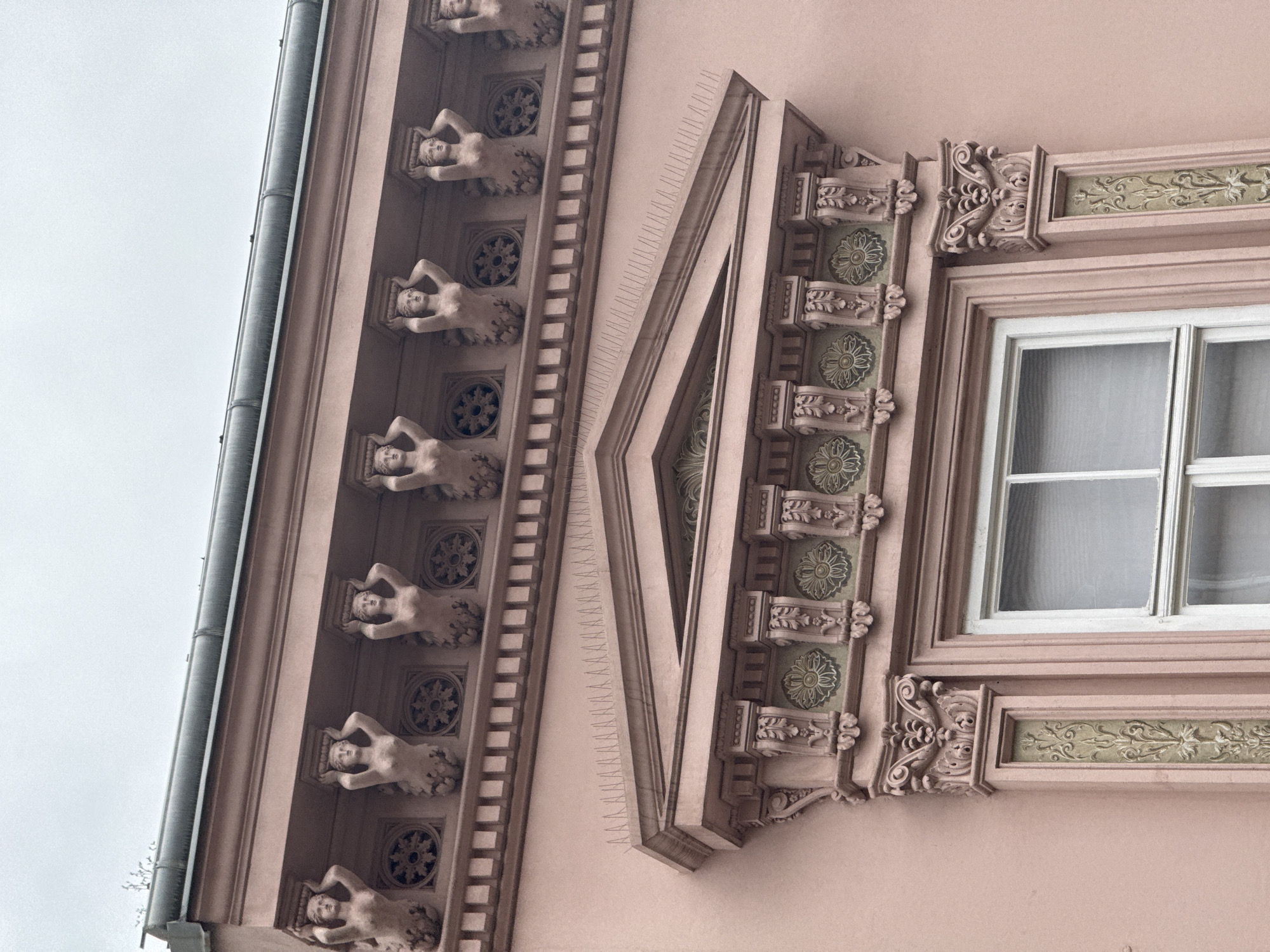 A low-angle medium shot depicts the corner of a building in Zagreb, Croatia. The building is pink, and the exterior is decorated with ornate architectural details. A vertical line of six female statues supports the structure, each figure reclining with hands positioned behind the head. Between each statue, a decorative floral motif is featured. To the right of the statues, a series of parallel lines run vertically along the building's corner. The corner is decorated with triangular motifs and floral patterns. A window is situated to the right, framed by additional ornamentation and elaborate carvings. The sky, visible at the upper left of the image, appears overcast. A gutter runs vertically along the building's edge, parallel to the line of statues.