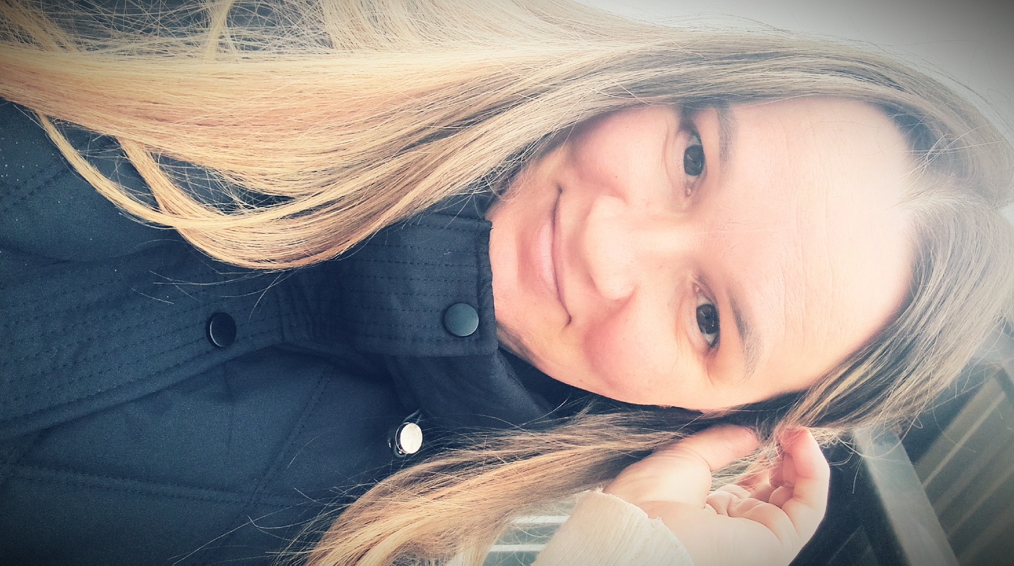 A woman with long blonde hair is seen from a close angle, wearing a dark jacket with buttons. She is smiling slightly and looking directly at the camera. Her right hand is near her hair. The location cannot be determined from the image.