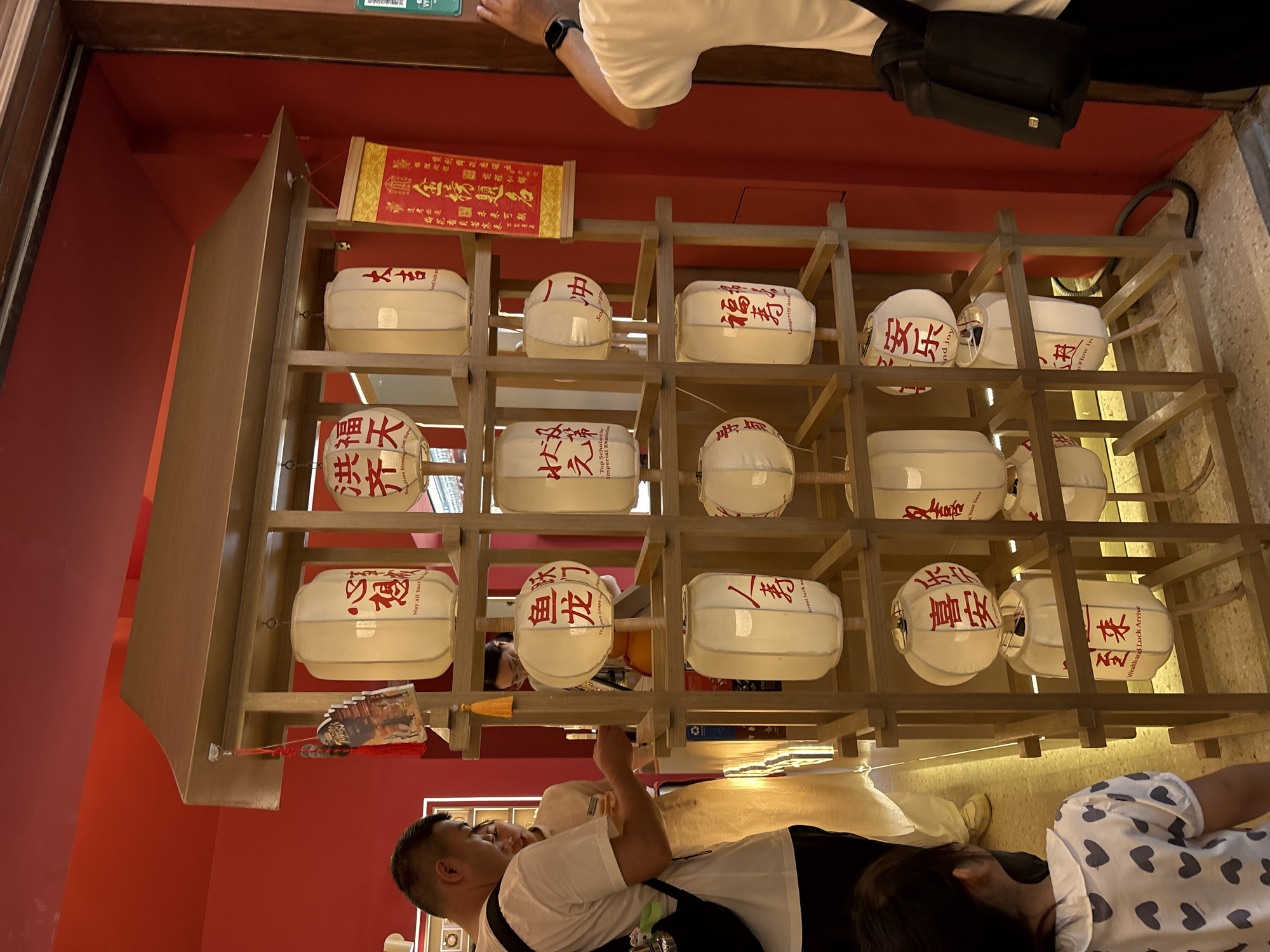 The image captures a display of traditional Chinese lanterns in Shanghai, China. The lanterns, made of white paper with red Chinese characters, are arranged in a wooden shelving unit. Some lanterns have English translations below the Chinese text, such as "May All Your..." and "Longevity and...". In the foreground, a young girl with dark hair, wearing a white dress with gray hearts, looks towards the lanterns. To the left, a man with short dark hair, wearing a white t-shirt and black pants, faces the display. Another woman in a light-colored dress is visible behind him, also looking at the lanterns. The background features a bright red wall, suggesting an indoor setting like a shop or cultural establishment. A yellow scroll with Chinese calligraphy hangs on the right side of the display. The overall scene is well-lit, highlighting the vibrant red and white colors.