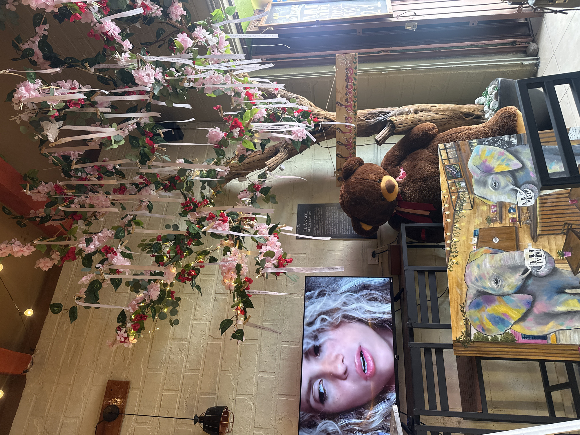 The image captures an indoor scene at a cafe in San Andrés Cholula, Mexico. A large, artificial tree decorated with pink and red artificial flowers and numerous white ribbons hangs from the ceiling. Some ribbons have handwritten messages on them. A sign with painted text "El Árbol de los deseos" (The Tree of Wishes) is attached to the tree trunk. To the right of the tree, a large stuffed teddy bear is seated, wearing a red and white striped scarf. In the foreground, a television screen displays a close-up of a woman's face. To the left of the TV, a table with a black metal frame supports the television. Below the TV, two framed artworks lean against the table; one depicts an elephant with colorful accents holding a mug. The walls are made of light-colored brick. String lights are visible above the tree. A doorway is visible in the background to the right.
