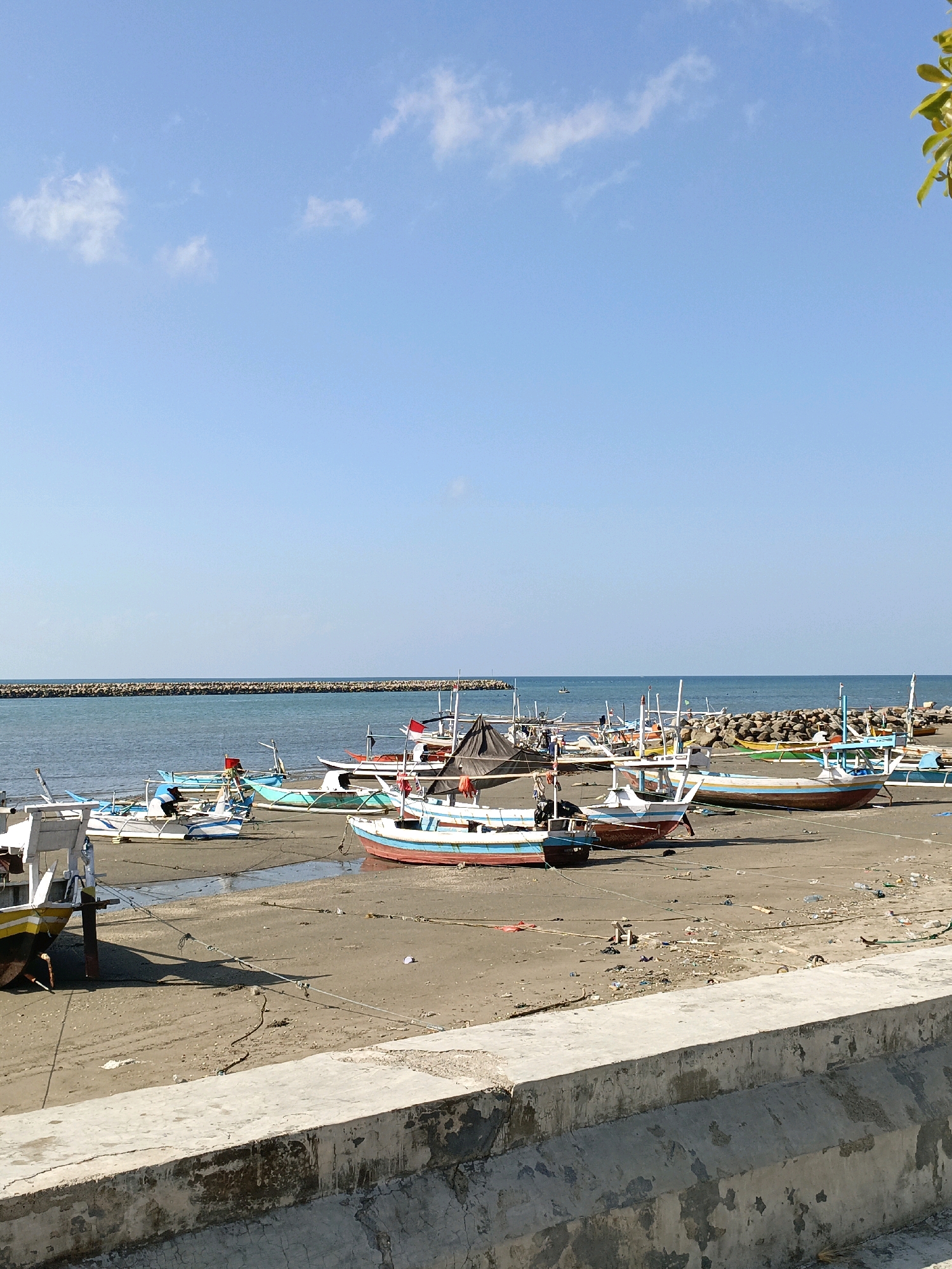 A wide shot shows a beach in Bulukumba, Indonesia, with numerous colorful fishing boats resting on the sand near the water. A long, rocky breakwater stretches across the horizon. The sky is a clear, pale blue with a few wispy clouds. The sand is littered with debris, including plastic bottles and other trash. In the foreground, a concrete wall runs along the edge of the beach.