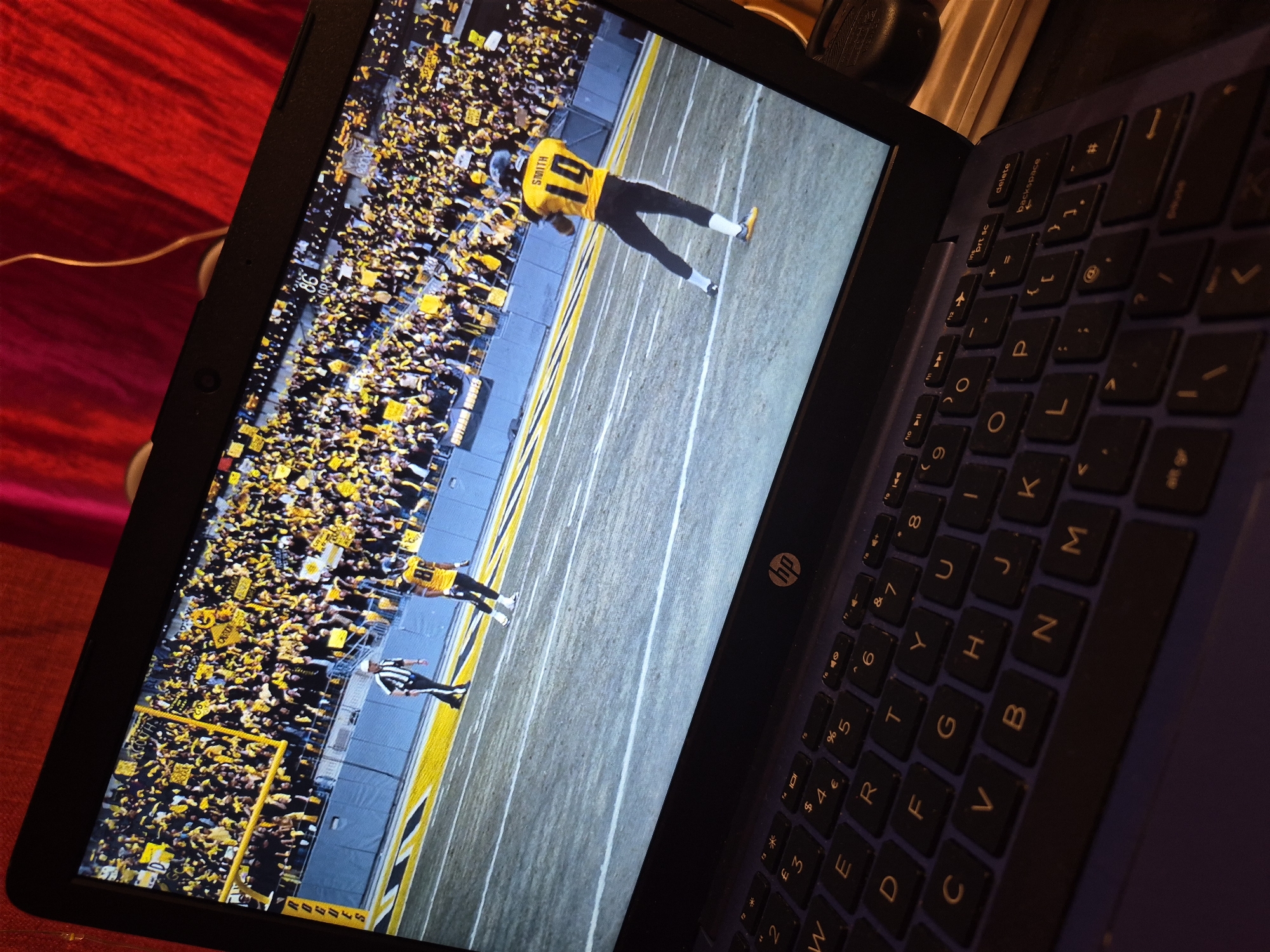 A laptop screen displays a live football game, featuring players in yellow and black uniforms on a field with yellow end zones. A crowd of spectators, predominantly wearing yellow, fills the stands. The laptop itself is positioned on a dark surface, with its keyboard visible in the foreground. The background is a textured red fabric. The image does not provide any information about the location in the city of Sandwell, United Kingdom.